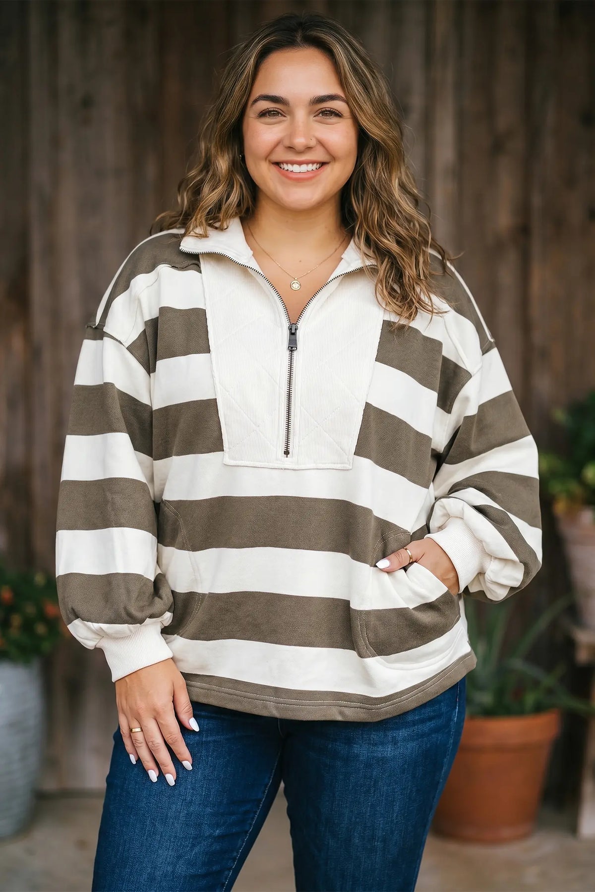 A woman wearing a brown and off-white horizontally striped quarter-zip pullover sweatshirt with pockets, paired with blue jeans, standing in front of a rustic wooden fence.