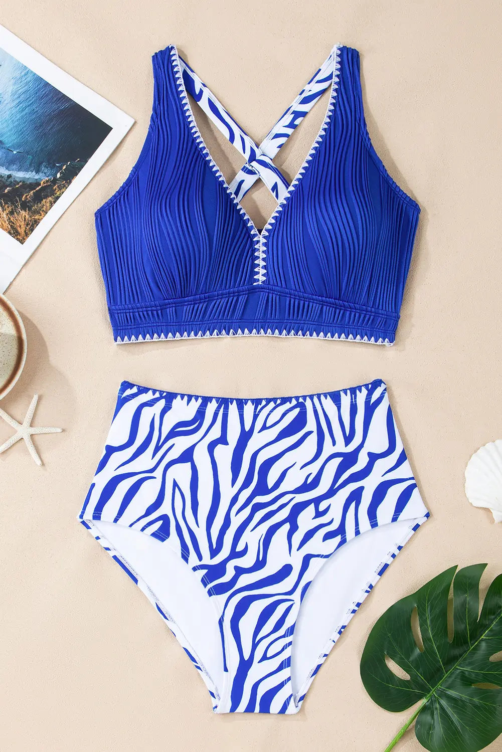 Blue and white patterned bikini set on a beige surface with a leaf and photo frame.