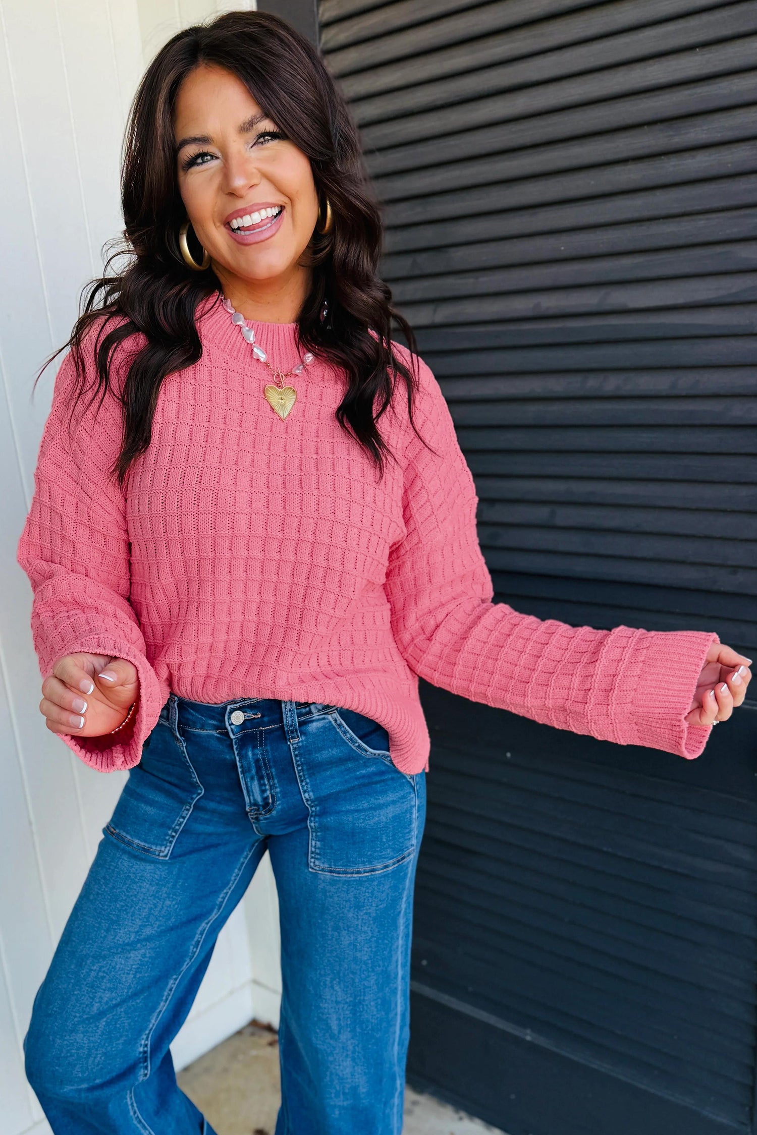 A woman wearing a pink textured knit sweater and blue jeans, smiling and posing in front of a dark gray slatted wall.