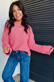 A woman wearing a pink textured knit sweater and blue jeans, smiling and posing in front of a dark gray slatted wall.