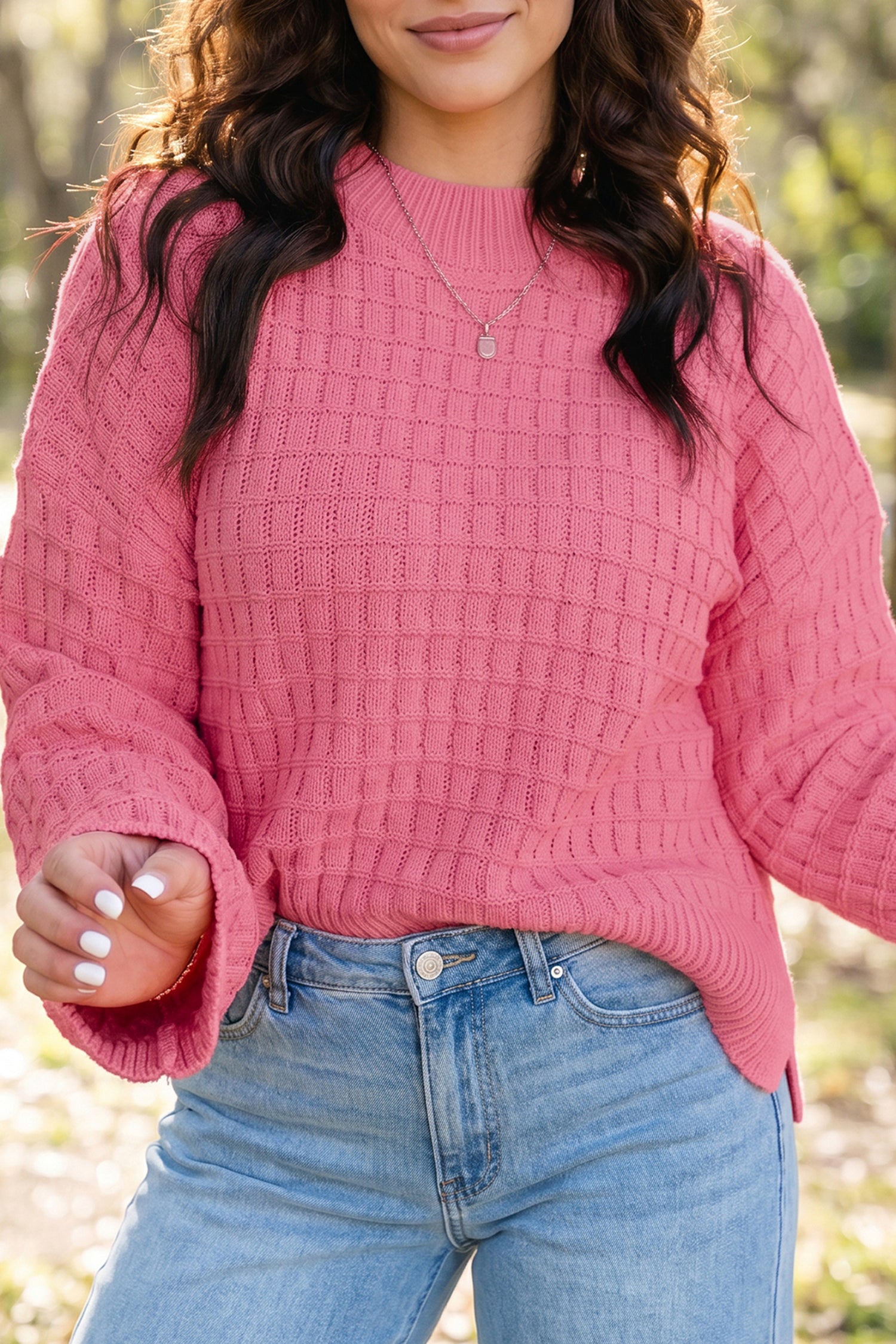 A woman wearing a pink textured knit sweater with a crew neck and flared bell sleeves, paired with light blue jeans, photographed outdoors against a blurred natural background.