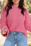 A woman wearing a pink textured knit sweater with a crew neck and flared bell sleeves, paired with light blue jeans, photographed outdoors against a blurred natural background.