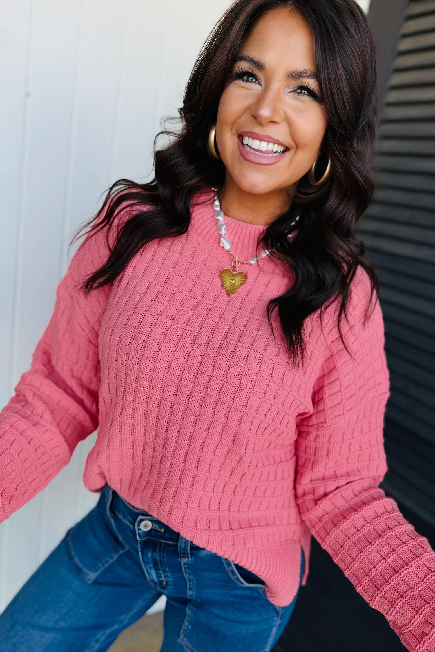 A smiling woman wearing a pink ribbed knit sweater and blue jeans, photographed against a light wall and dark slatted panel.