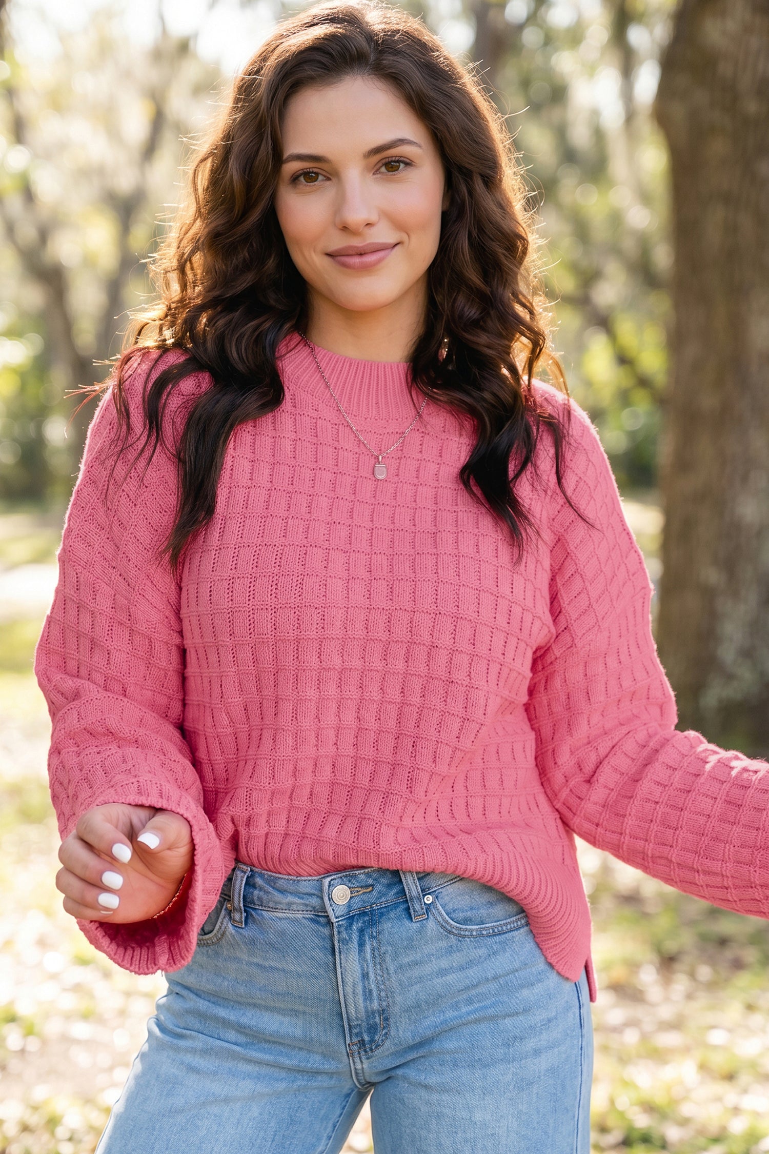 A woman wearing a pink cable-knit long-sleeve sweater and light blue jeans, standing outdoors in a park-like setting with blurred trees in the background.