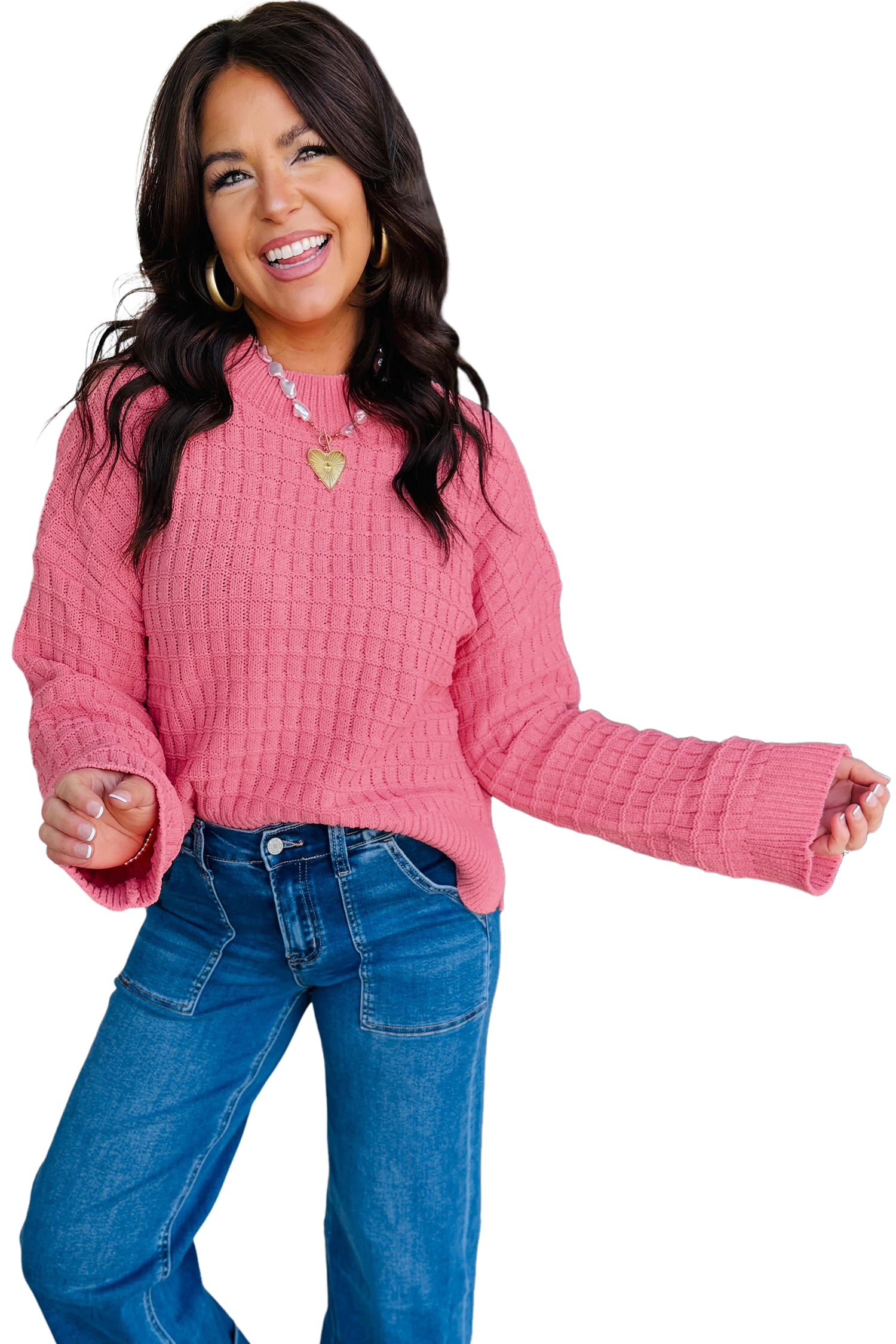 A woman modeling a pink textured knit sweater with long sleeves and a crew neck, paired with blue jeans, against a solid white background.