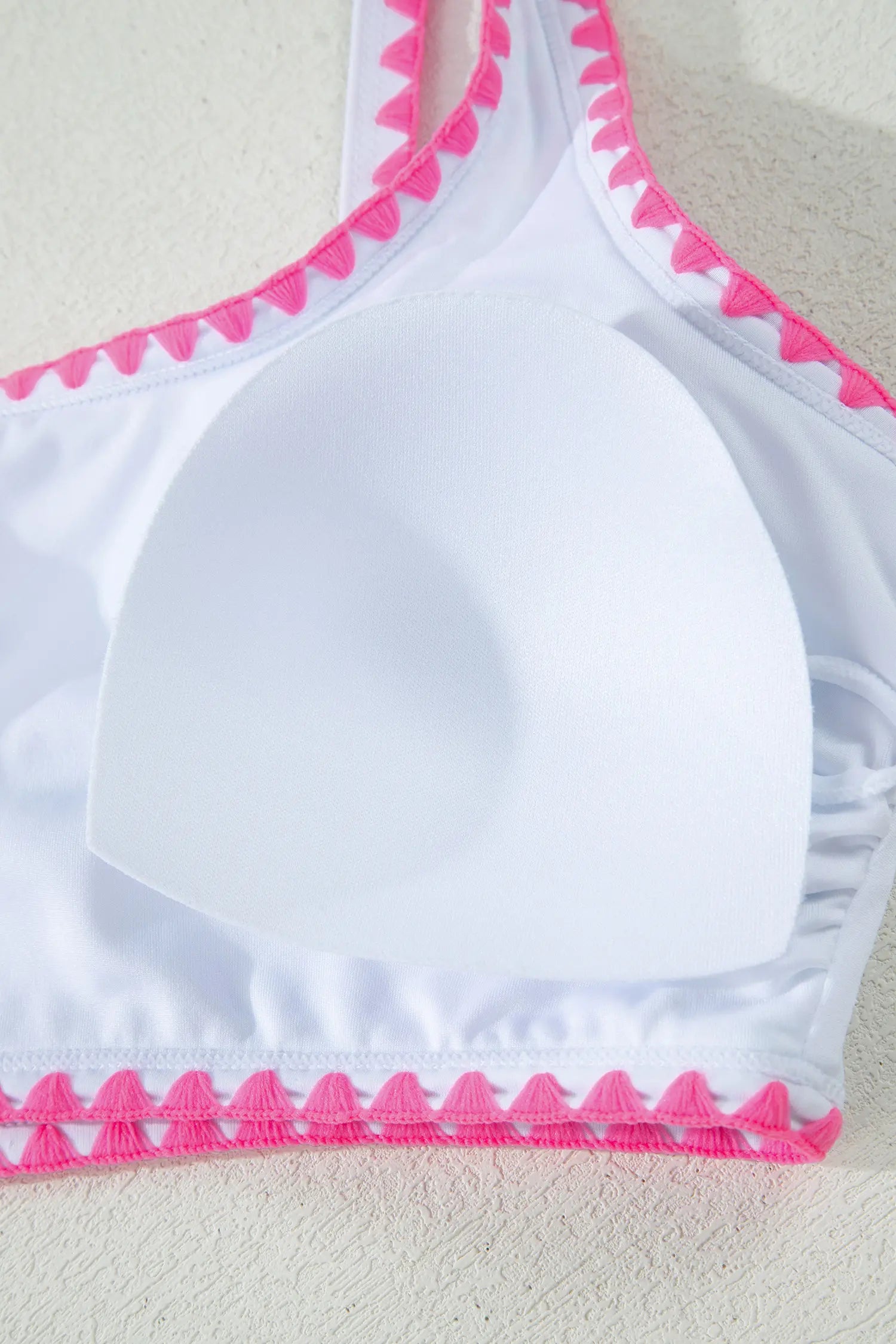 White bikini top with bright pink scalloped trim along the neckline and hem, shown close up on a light textured background.