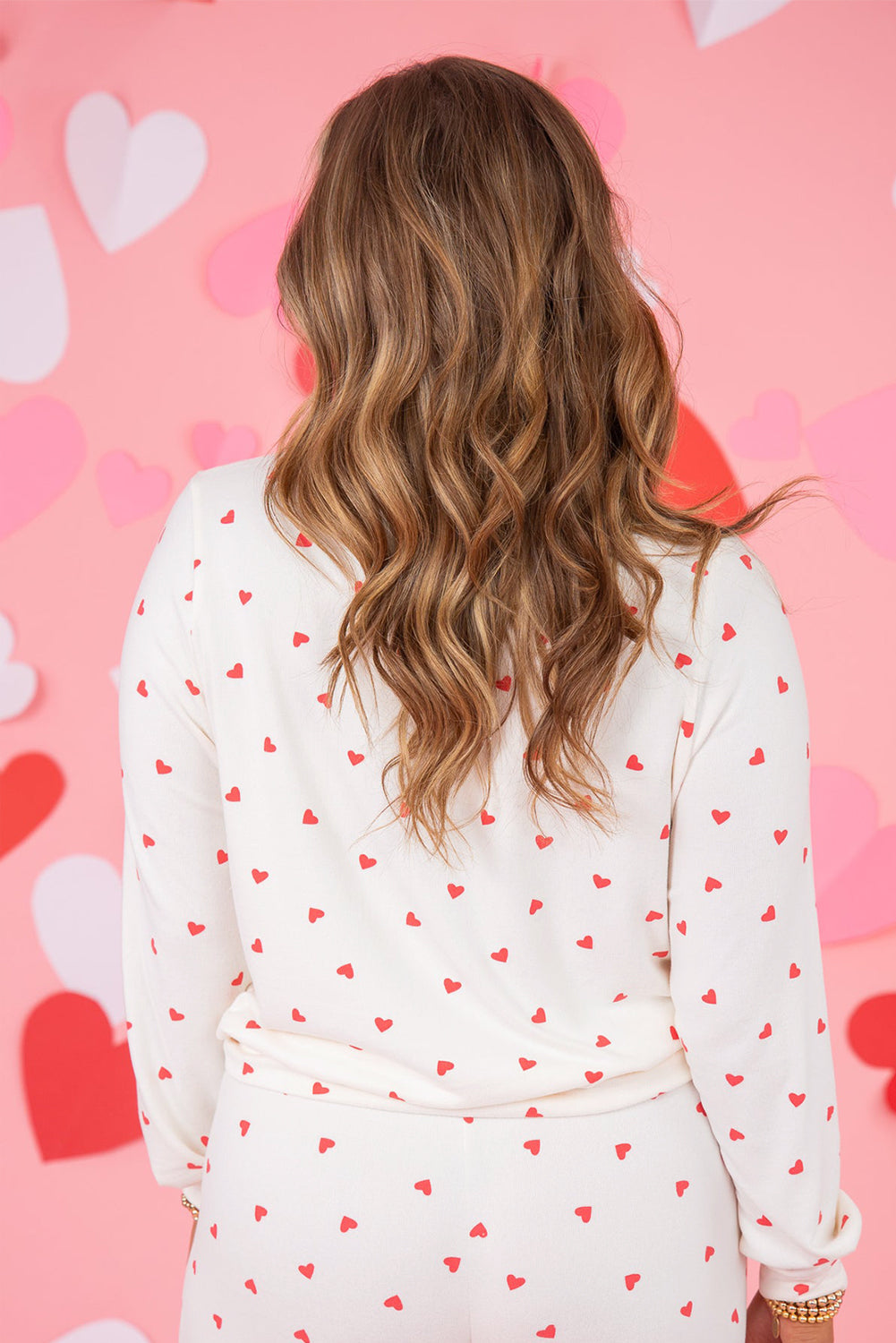 Person wearing a white long-sleeve top with red heart patterns against a pink background with heart shapes.