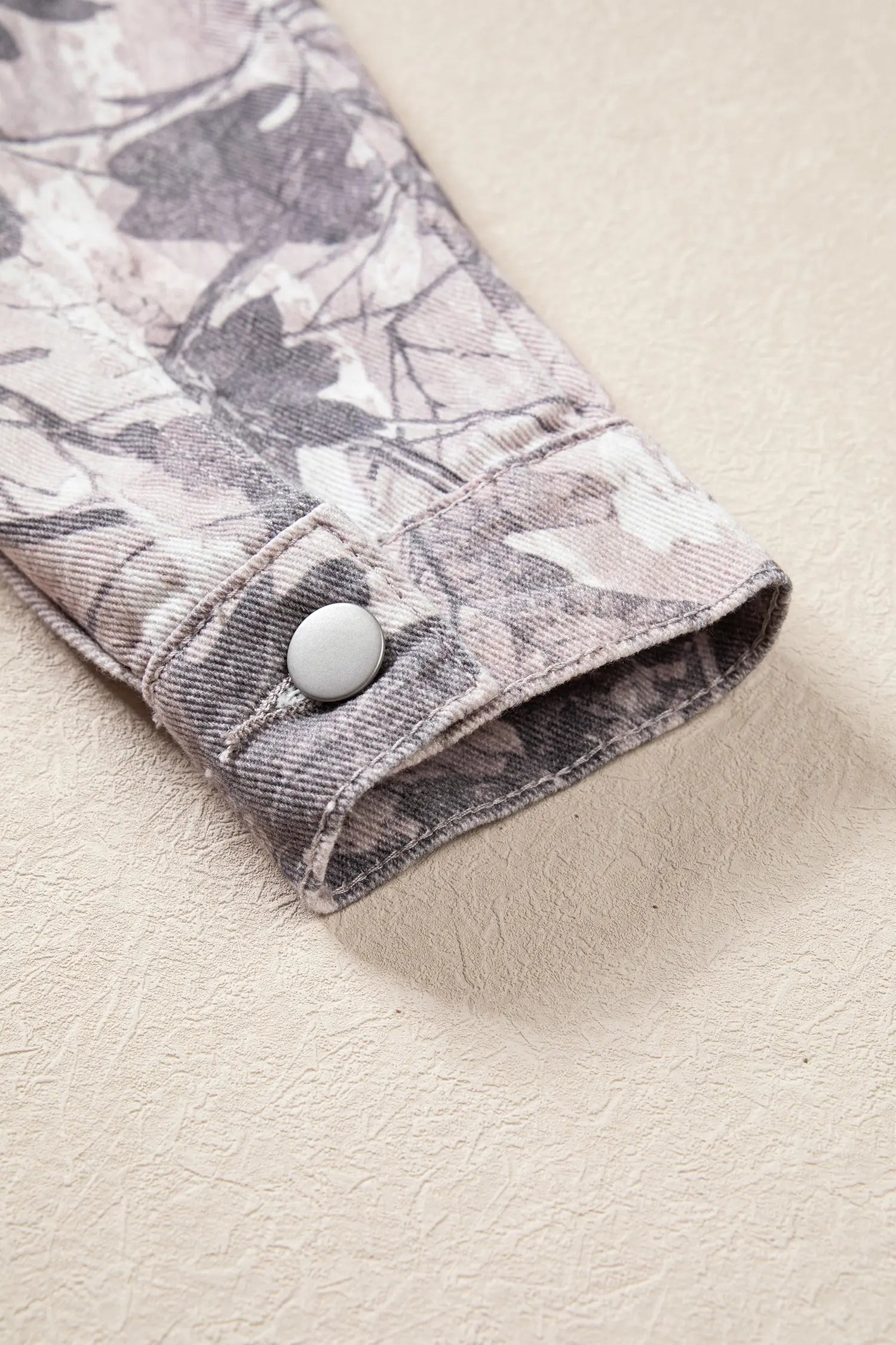 Close-up of denim jacket cuff with gray-beige leafy camouflage print and silver snap button.