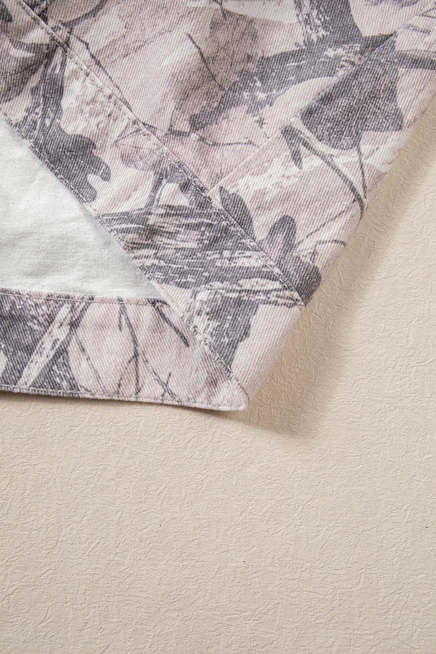 Folded garment with muted dusty pink, beige, and gray camouflage print on a textured beige surface.