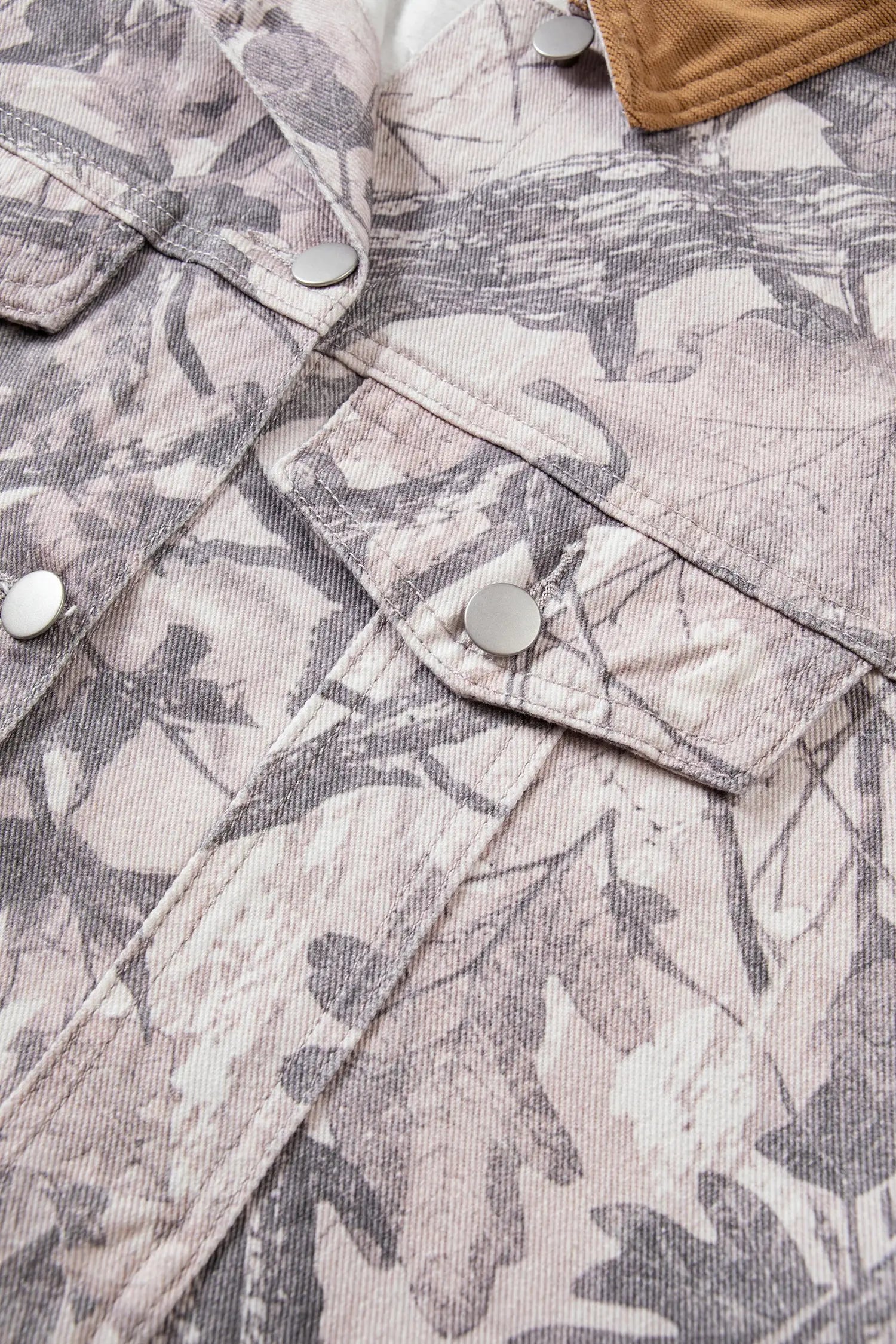 Close-up of light beige camouflage jacket fabric with dark gray leaf print and silver snap buttons.