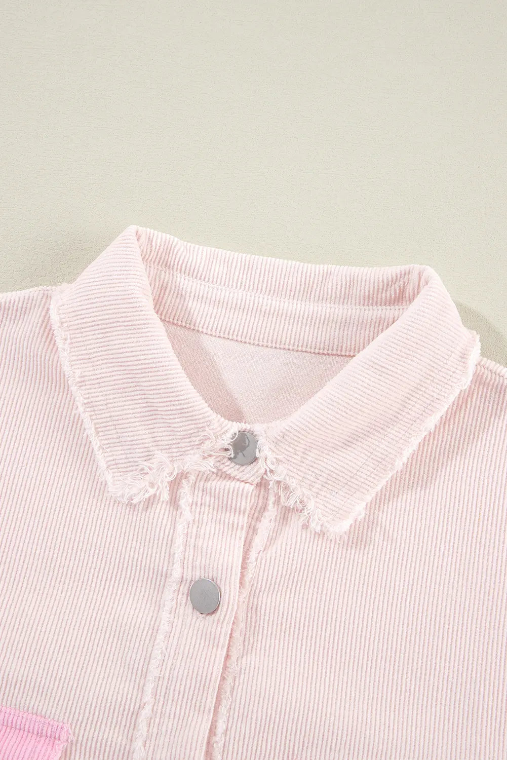 Close-up of a light pink corduroy collared shirt with a raw-edge collar and silver buttons, set against a neutral cream background.