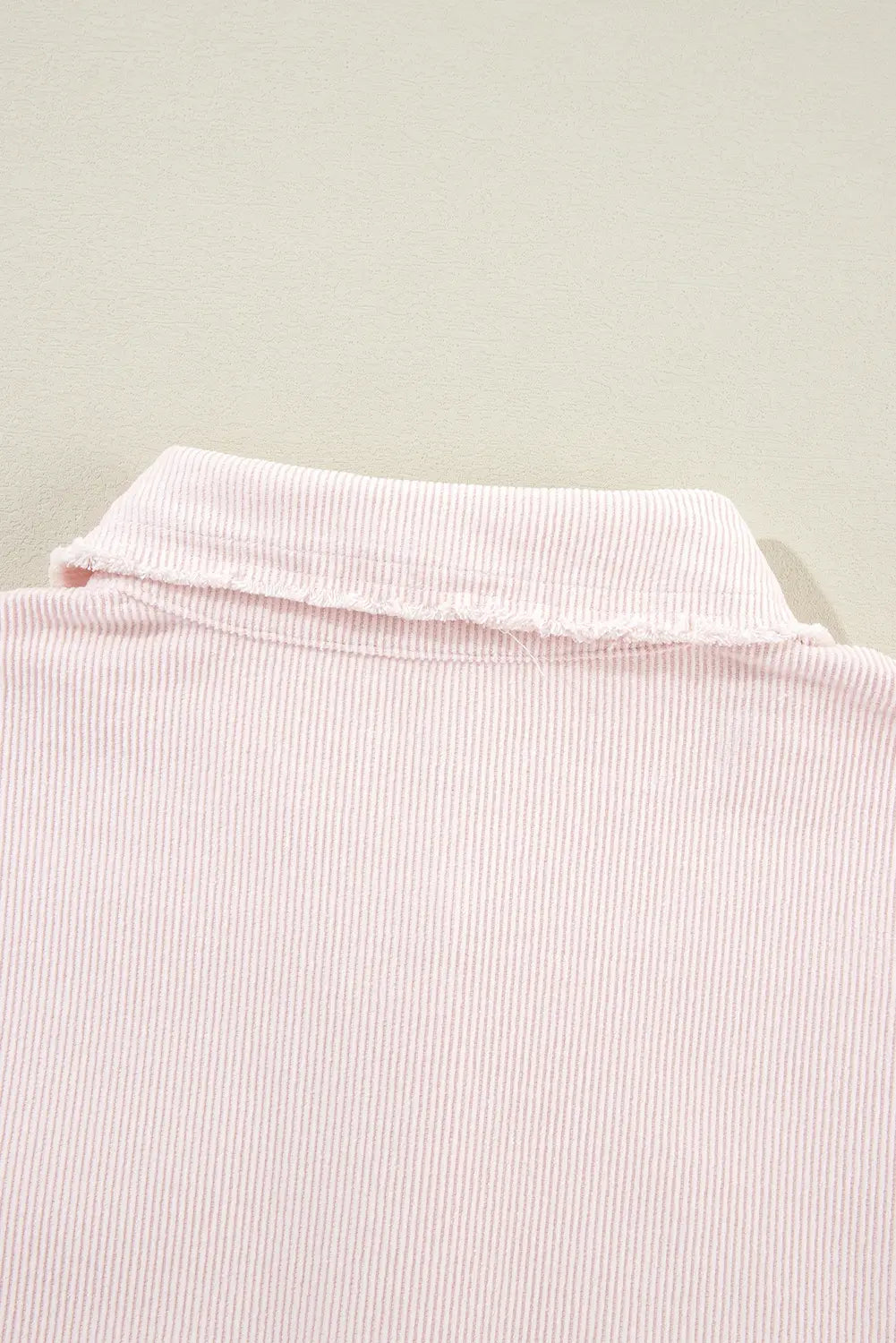 Close-up of a light pink ribbed knit garment, showing the collar and fine vertical stripe texture against a plain light beige background.