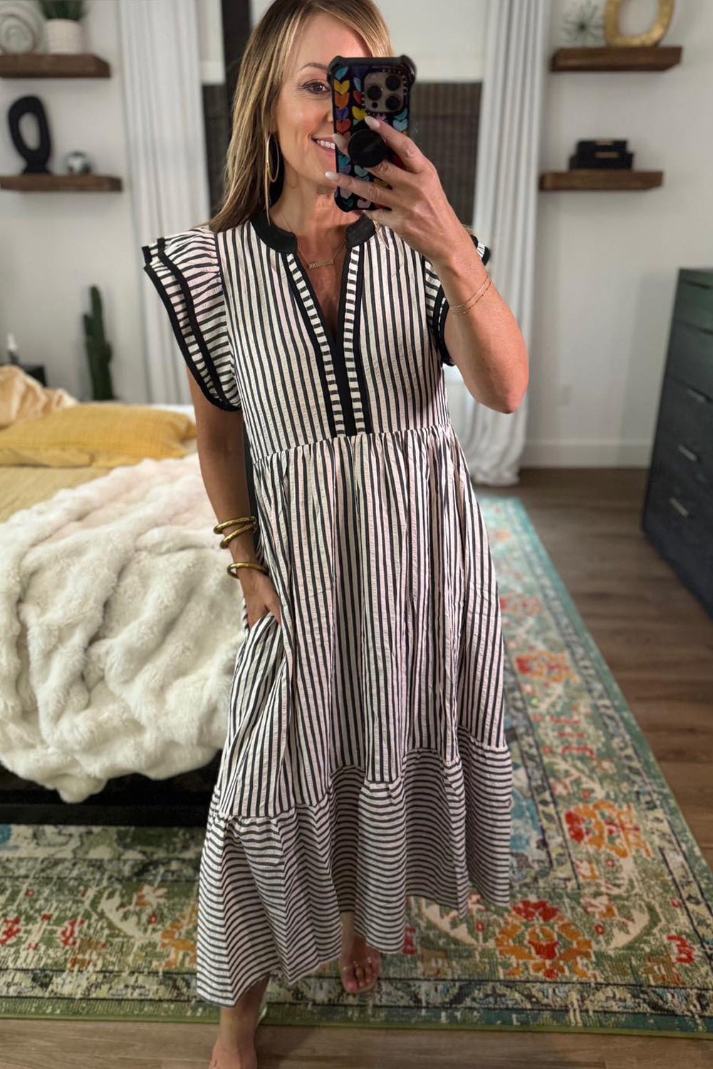 Woman taking a mirror selfie wearing a black and white striped dress in a room with a bed and shelves.