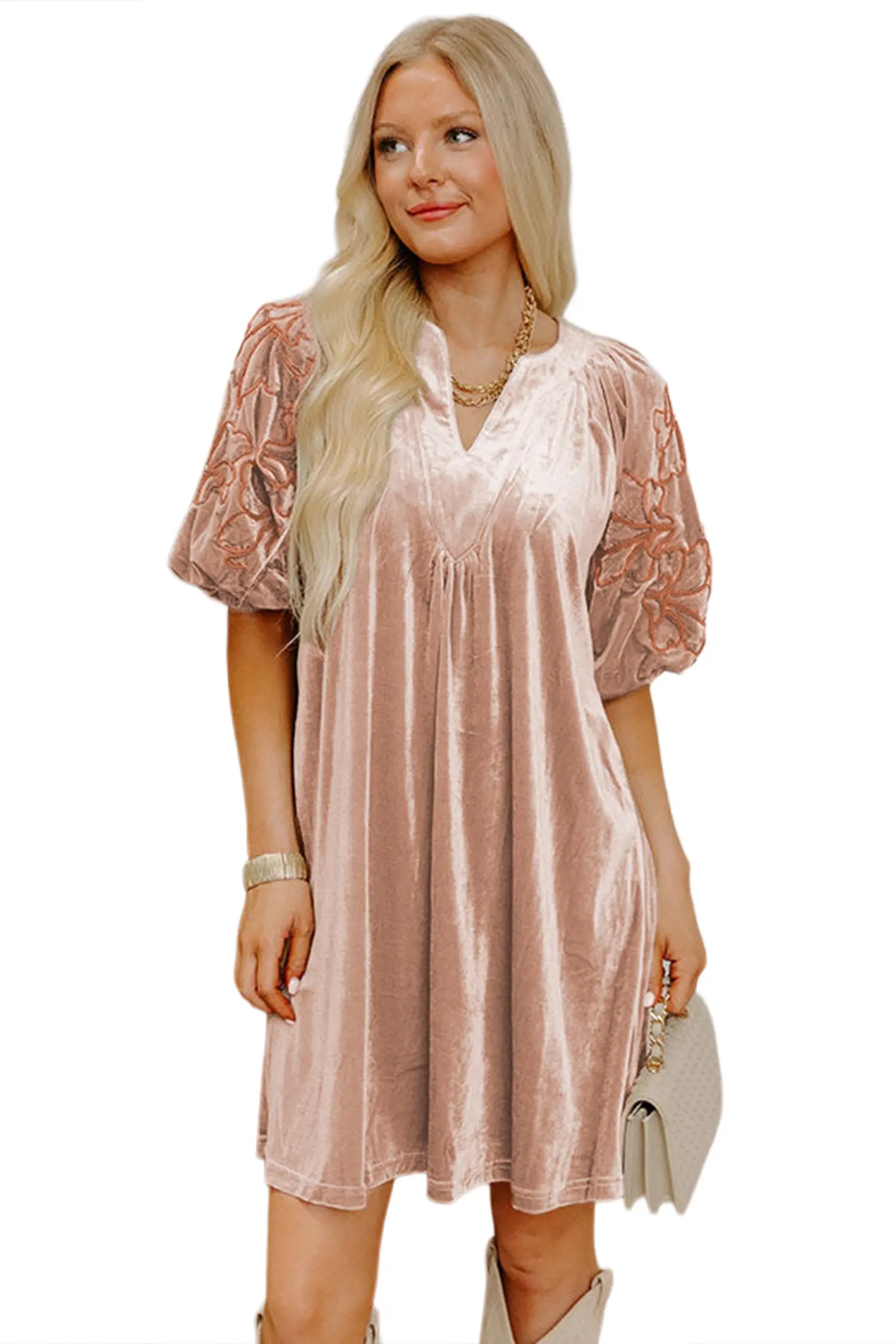 A woman modeling a short dusty pink velvet dress with sheer lace puff sleeves, holding a light beige clutch bag against a plain white background.