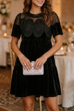 A woman wearing a black velvet and mesh mini dress, holding a silver patterned clutch bag in a softly lit indoor event setting.