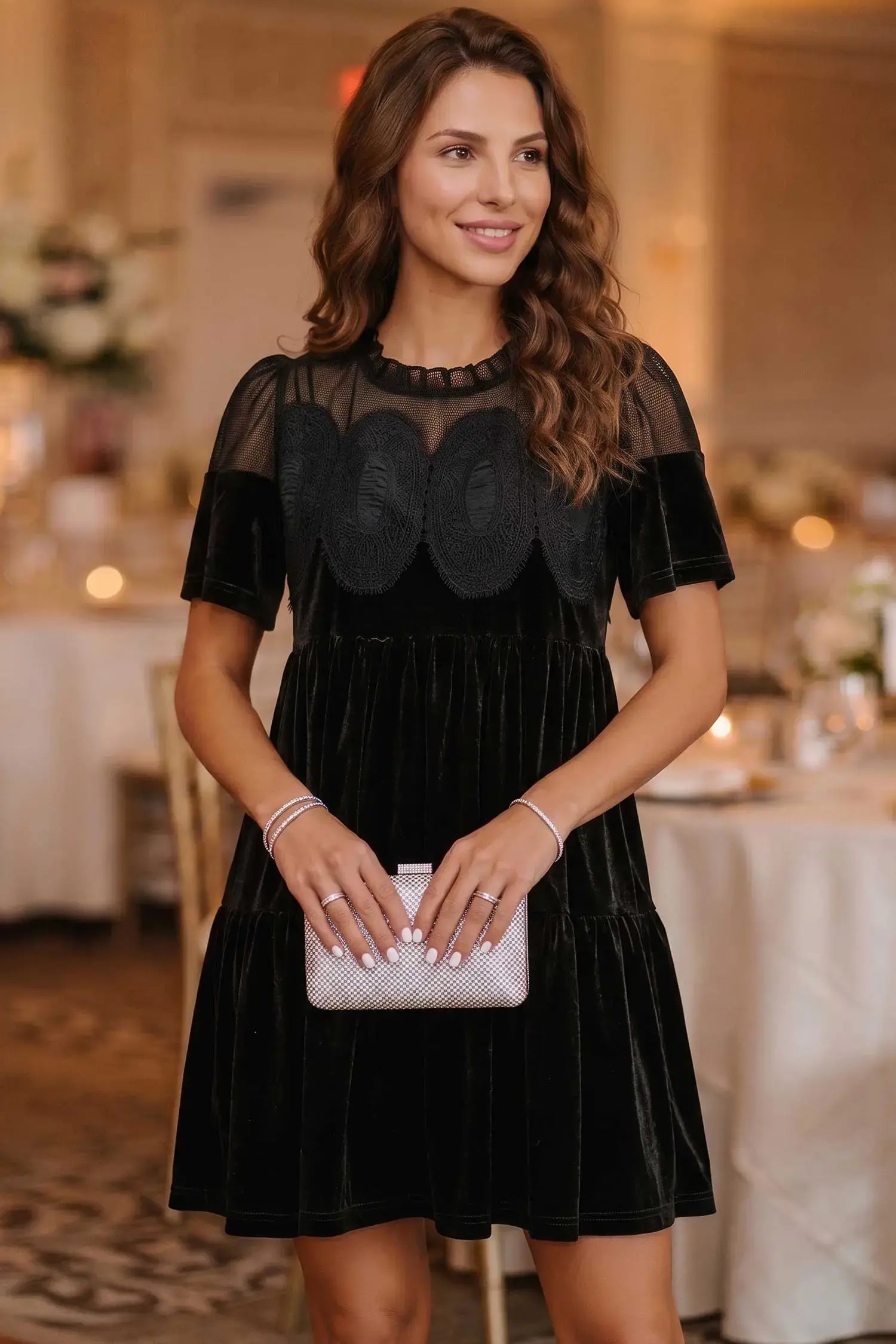 A woman wearing a black velvet tiered dress with sheer mesh short sleeves and a scalloped black overlay bodice, holding a silver clutch in a softly lit event venue.