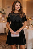 A woman wearing a black velvet tiered dress with sheer mesh short sleeves and a scalloped black overlay bodice, holding a silver clutch in a softly lit event venue.