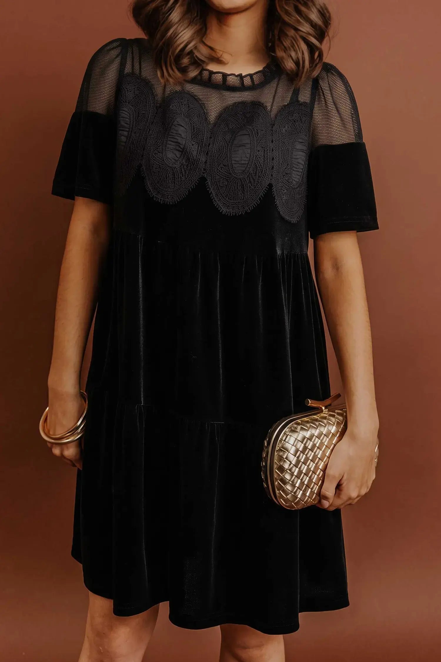 A woman wearing a black dress with sheer mesh short sleeves and a scalloped embroidered yoke, holding a small metallic clutch against a warm brown background.