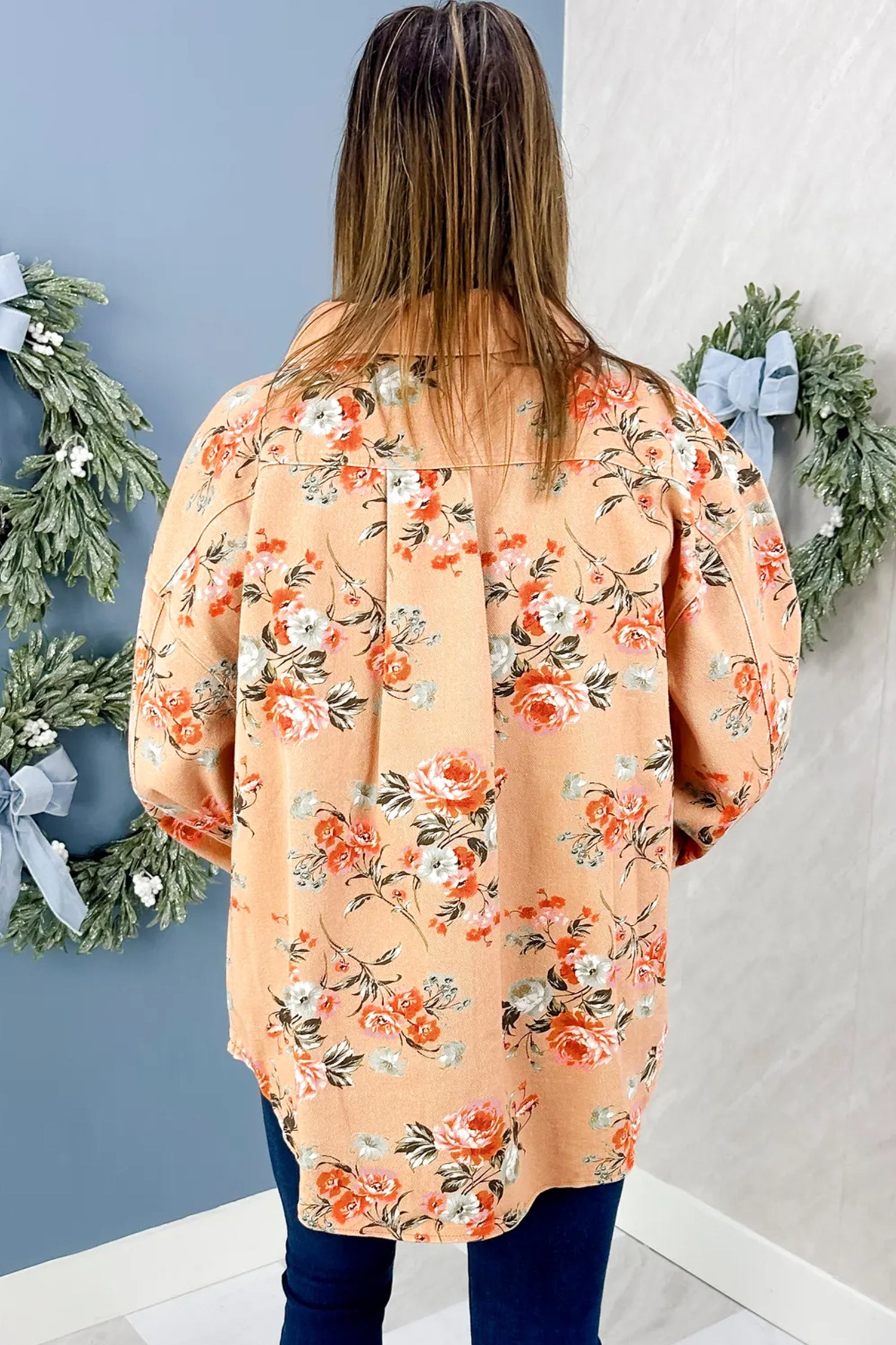 Woman in oversized peach floral blouse and dark blue jeans, viewed from behind indoors.