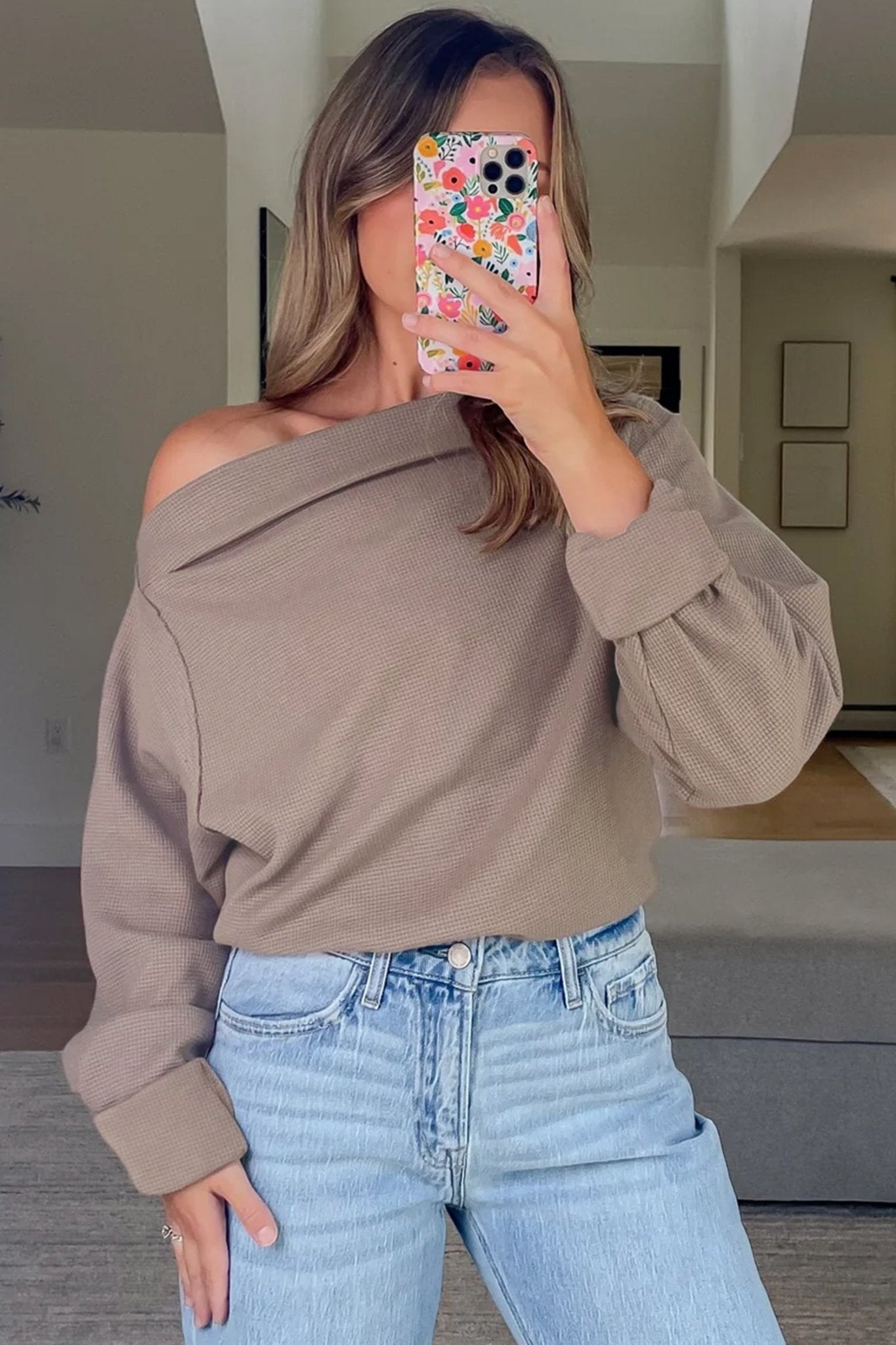 Woman wearing a beige off-shoulder sweater and blue jeans, taking a mirror selfie.