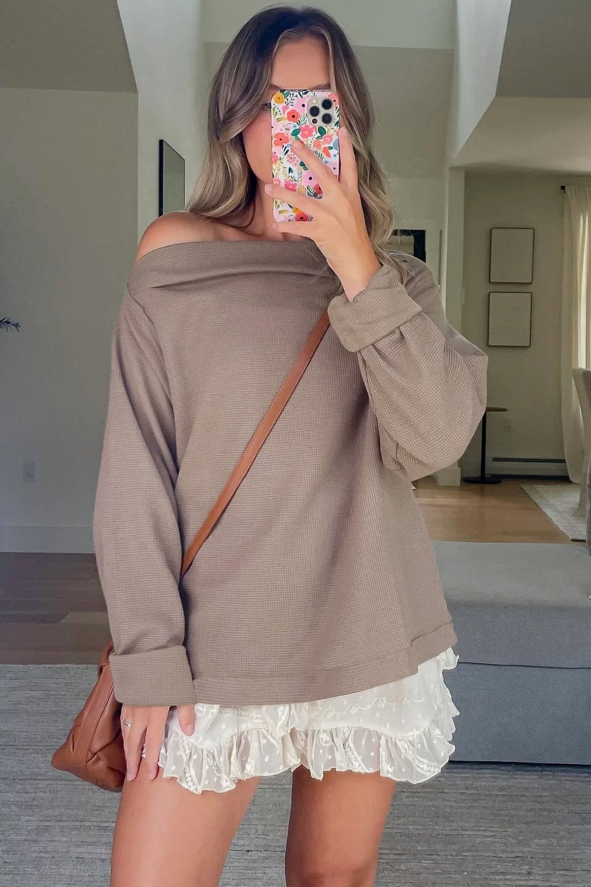 Woman wearing a brown off-shoulder sweater over a white lace dress, holding a phone with a floral case.