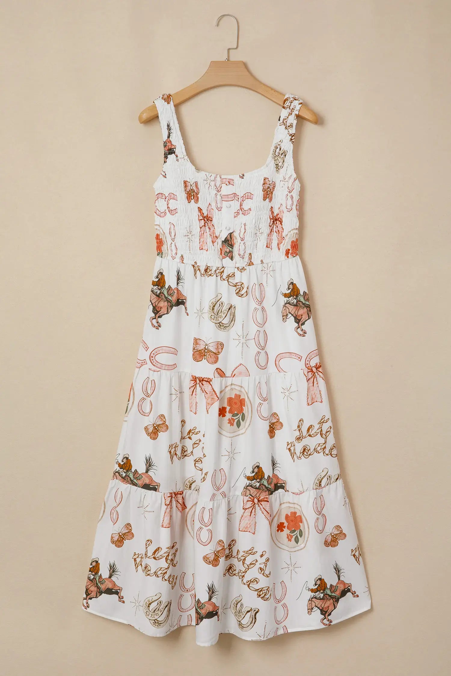 A white sleeveless sundress with a smocked bodice and tiered skirt, featuring an all-over whimsical print of fairies, bows, and autumn motifs in orange, brown, and beige, hanging on a wooden hanger against a solid beige wall.