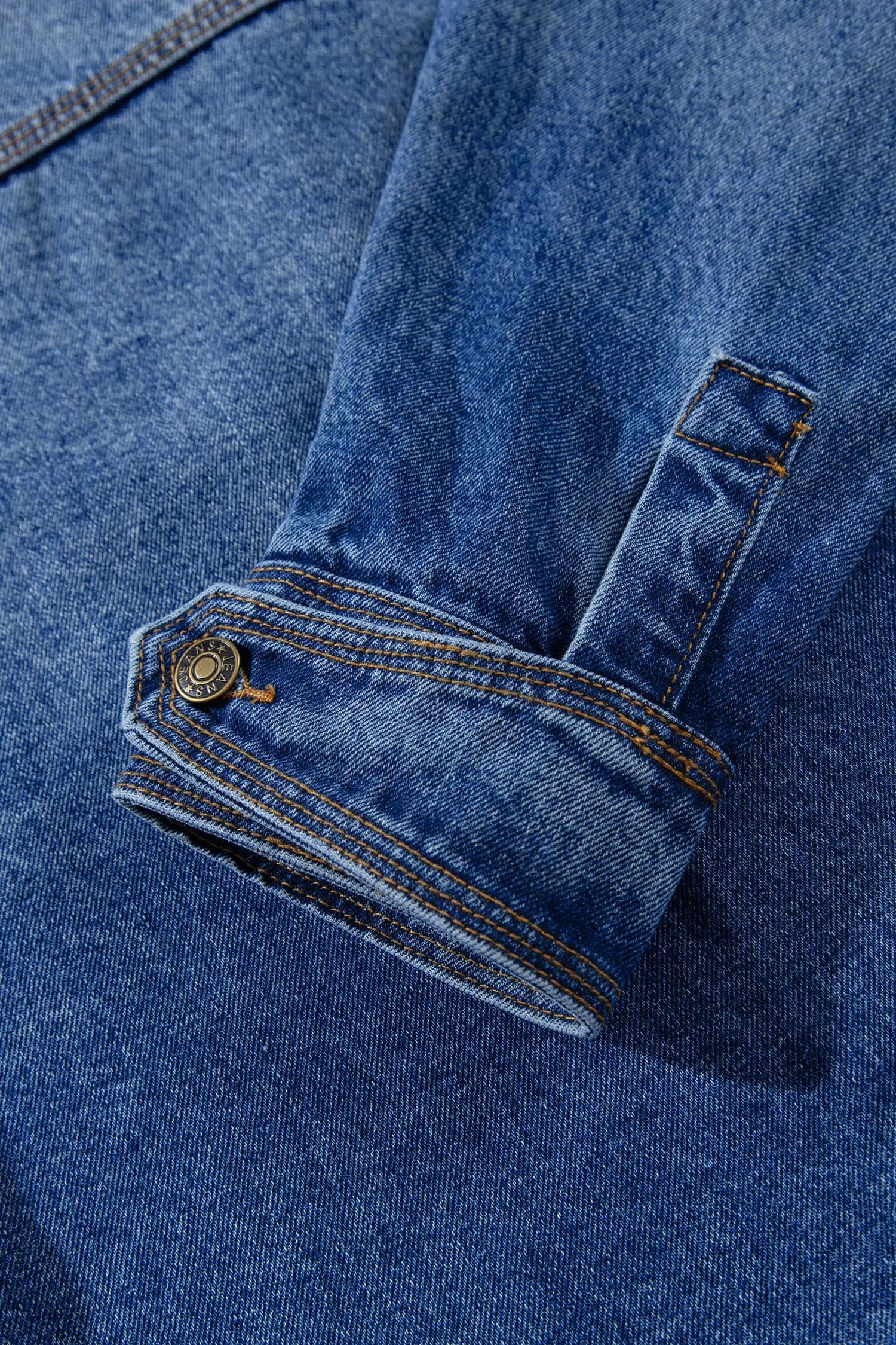 Close-up of blue denim garment with folded cuff, button tab, orange stitching