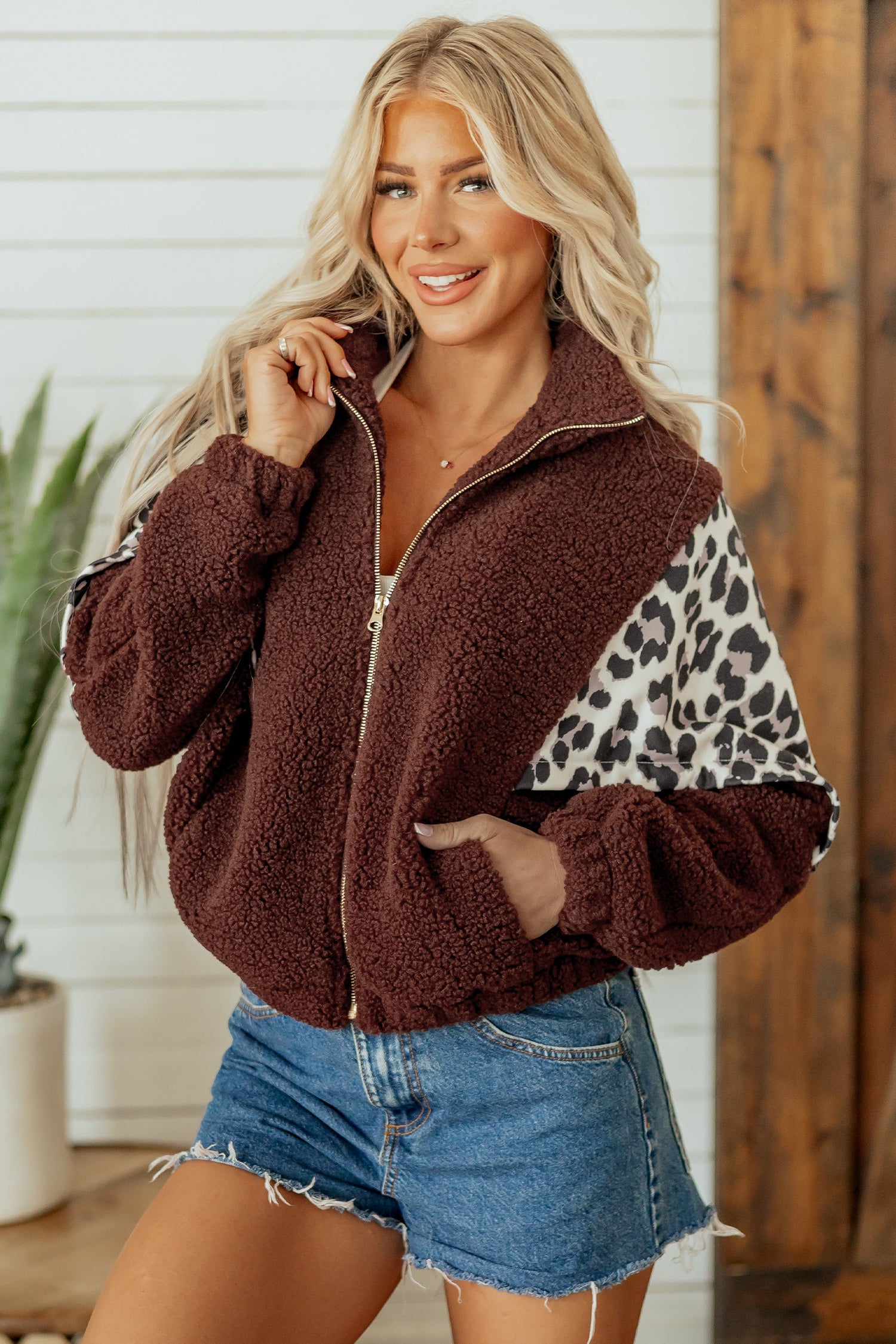 Woman in brown sherpa zip-up jacket with leopard panels and denim shorts.
