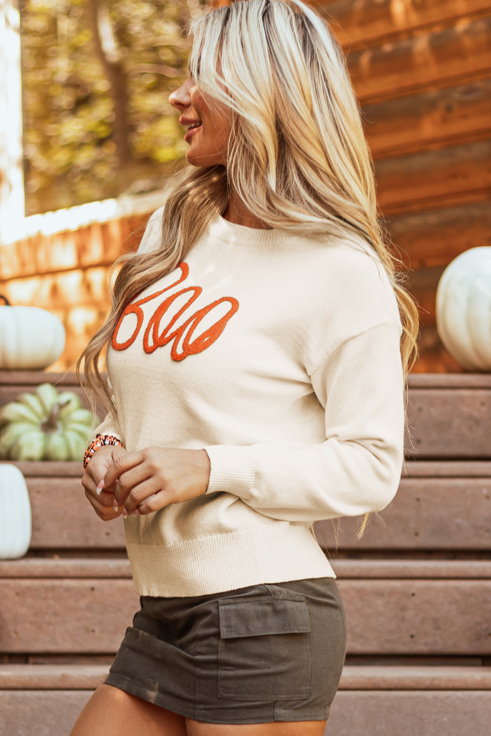 Woman wearing a white sweatshirt with red text, standing in front of pumpkins.
