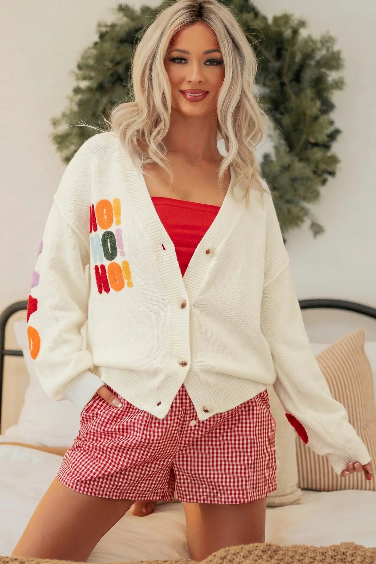A woman models a cream-colored knit cardigan with colorful 'Merry Merry' text graphics on the front and sleeve, worn over a red tube top and red-and-white gingham shorts in a bedroom setting.