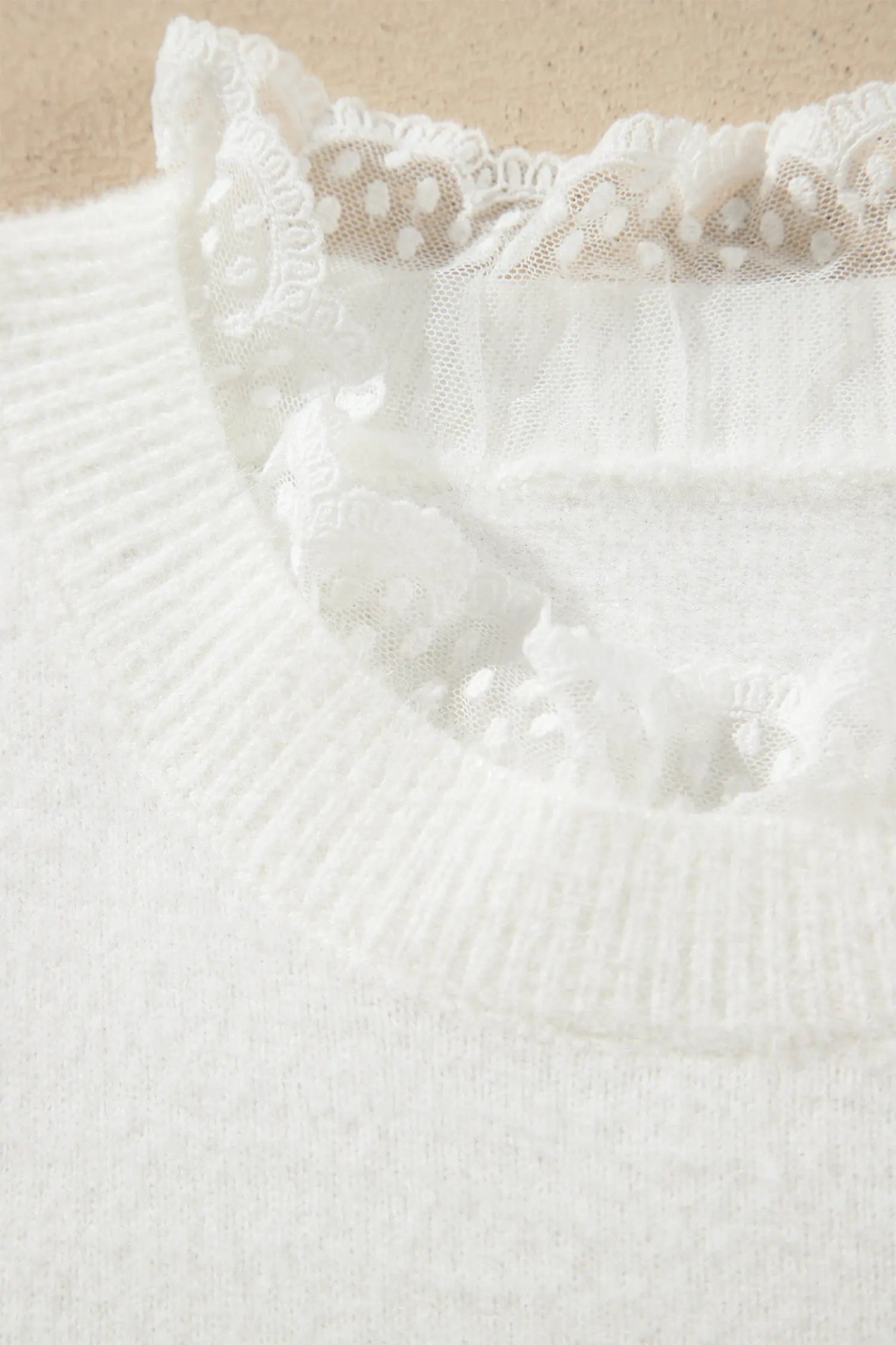 Close-up of a white knit sweater featuring a delicate white lace ruffle trim around the crew neckline, set against a neutral beige background.
