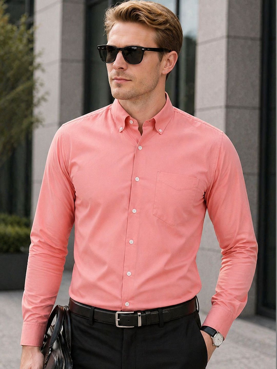 Man wearing a pink shirt and sunglasses standing against a building.