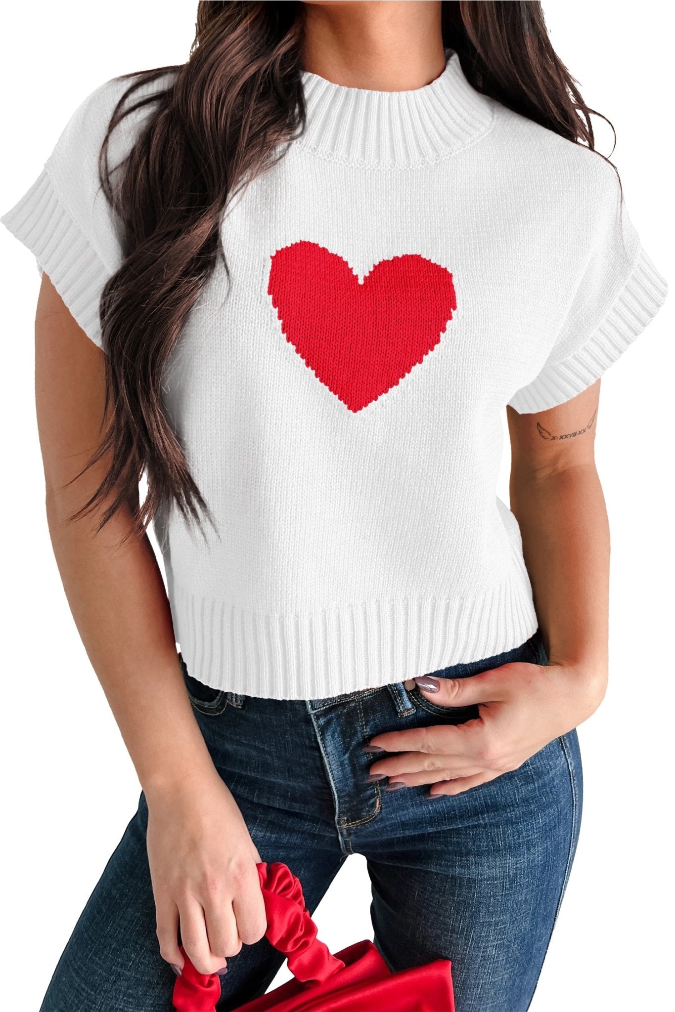 White sweater with a red heart design worn by a person, holding a red bag.