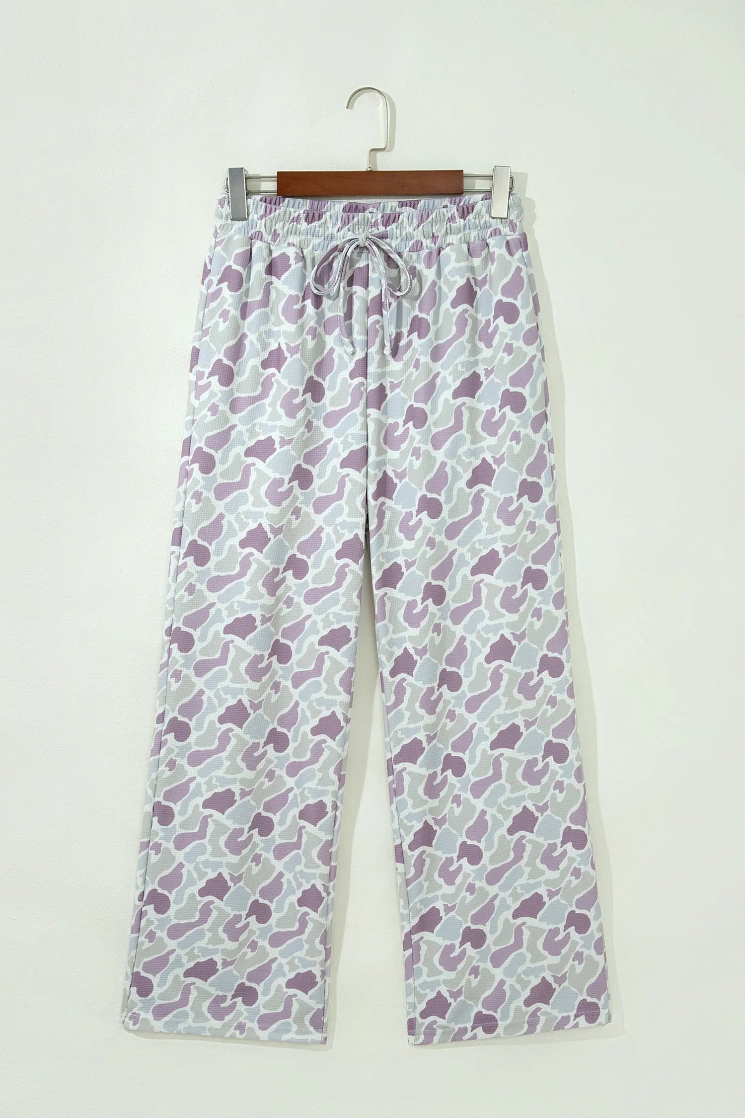 A pair of wide-leg lounge pants with a purple and white abstract pattern, hanging on a wooden hanger against a plain off-white wall.