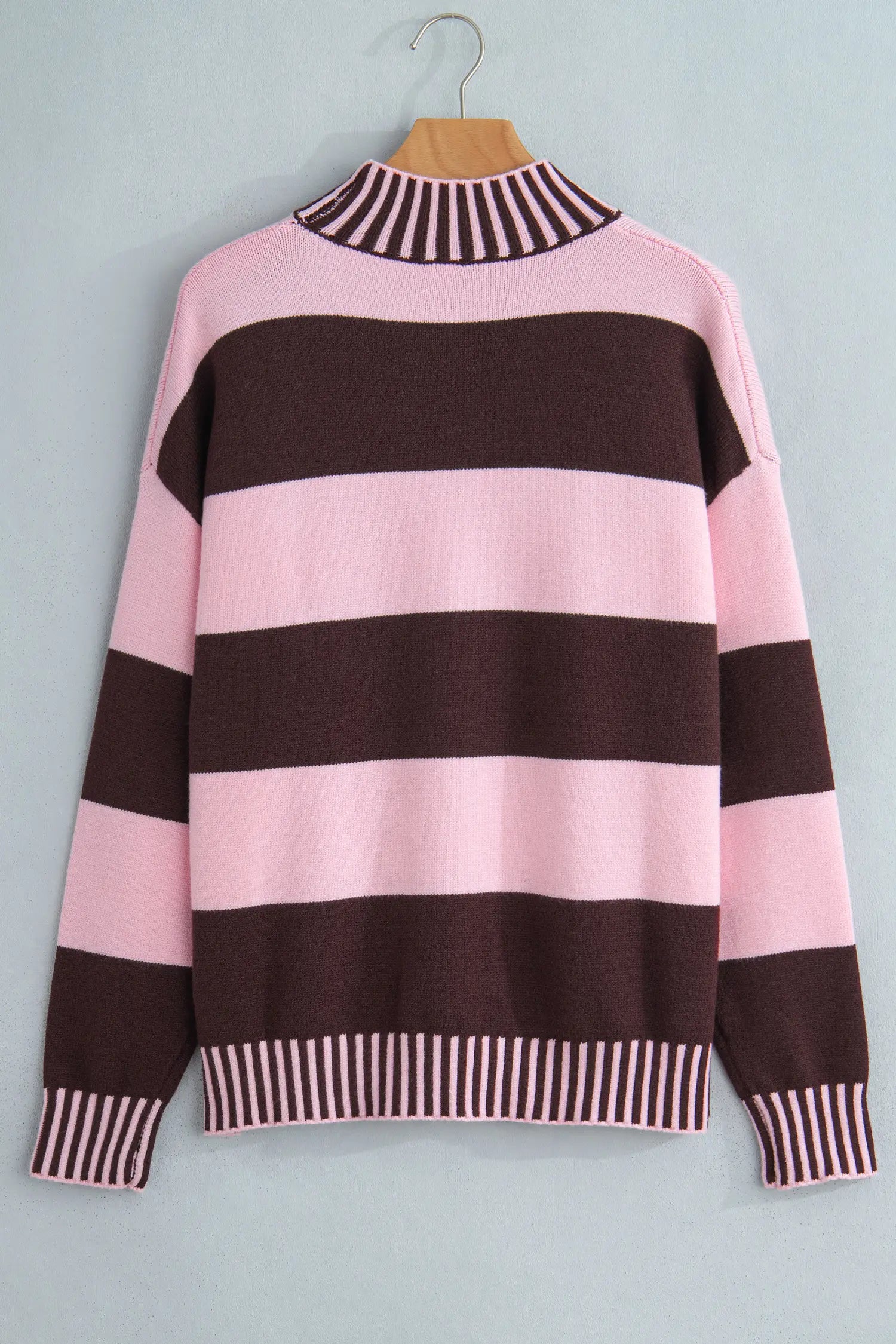 A pink and dark brown striped knit sweater with a mock neck, long sleeves, and striped ribbed cuffs and hem, hanging on a wooden hanger against a light gray wall.