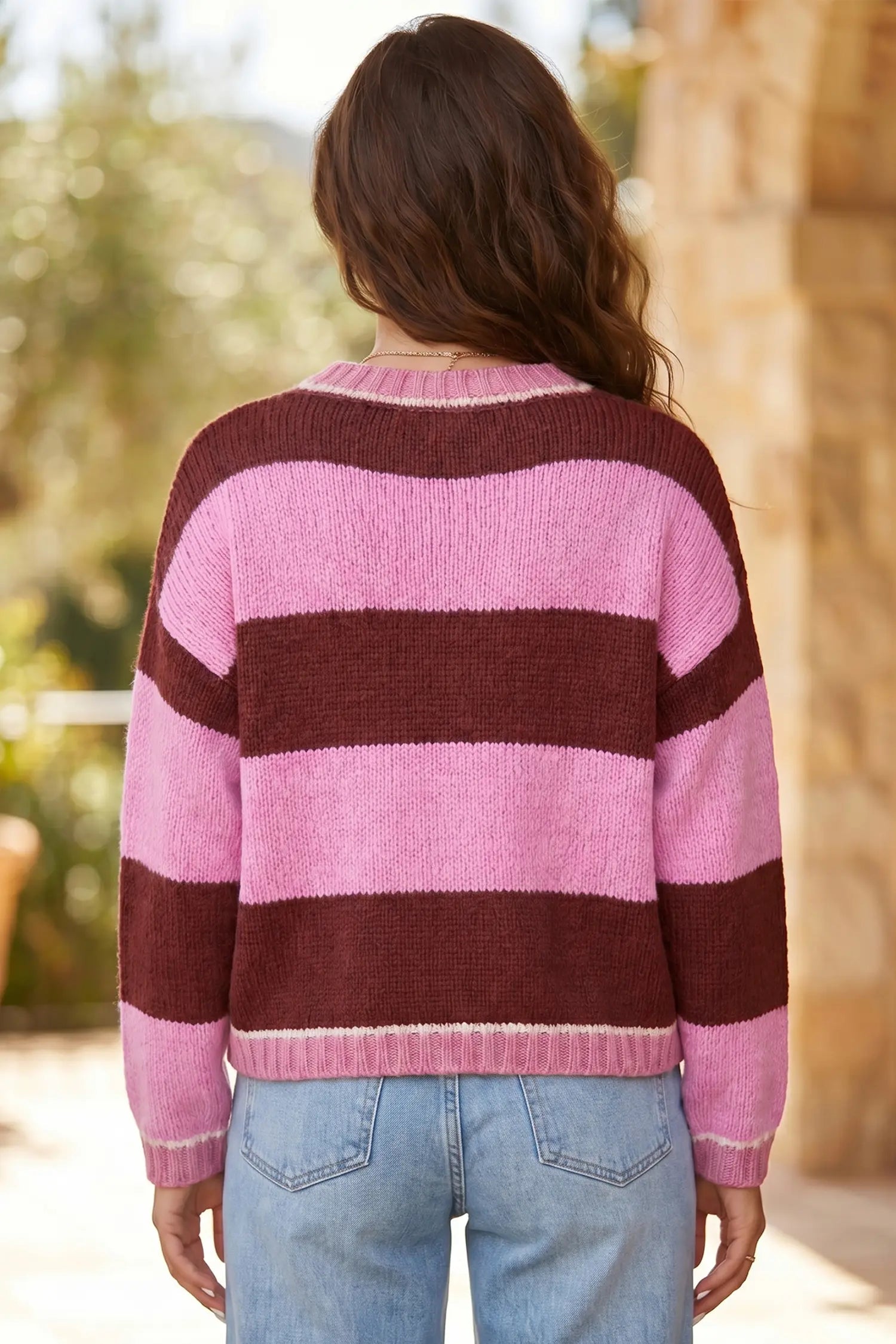 A woman viewed from behind wearing a fuzzy knit sweater with wide horizontal stripes in light pink and dark burgundy, paired with light blue jeans, set against a warm, blurred outdoor background.