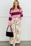 A woman stands indoors wearing a pink and burgundy striped knit sweater, wide-leg floral pants, and holding a dark burgundy handbag. The background is a white paneled wall with a gray floor.