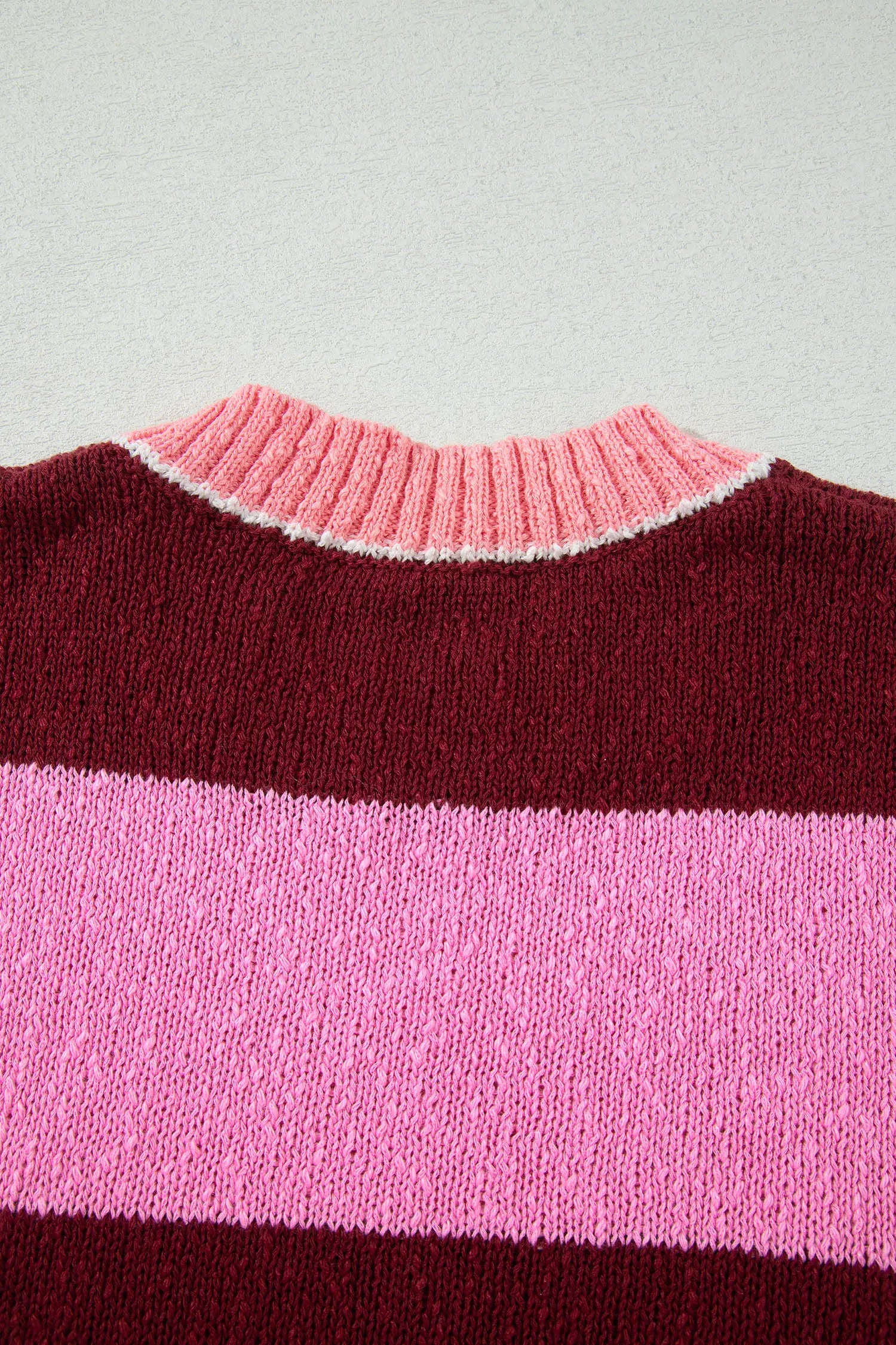 Close-up of a striped knit sweater showing a dark burgundy top section, a wide light pink middle stripe, and a thin white stripe separating the pink and burgundy sections, with a pink ribbed crew neck.