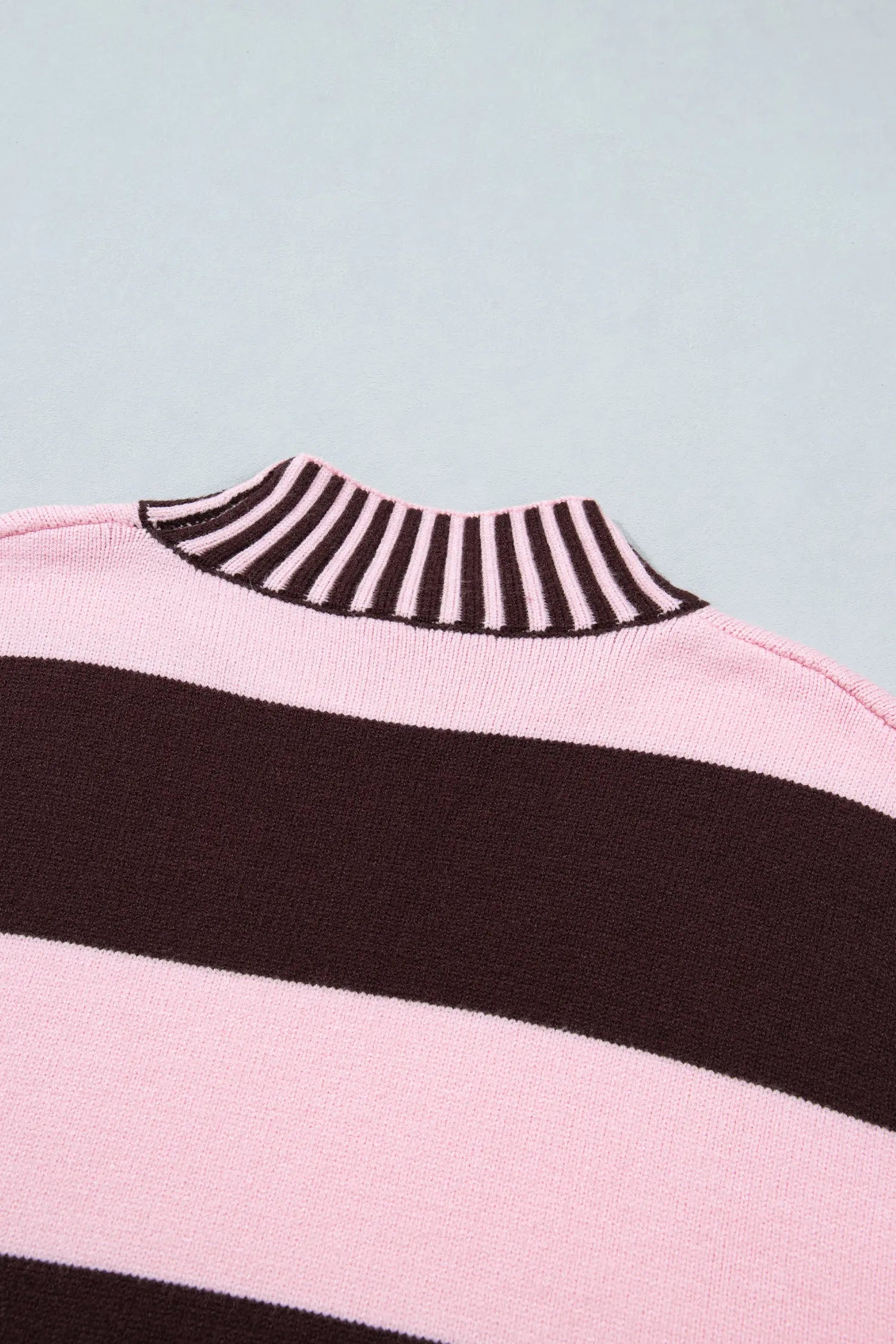 Close-up of a striped knit sweater showing a light pink body with wide dark brown horizontal stripes and a dark brown and light pink striped ribbed mock neck collar, set against a light blue-gray background.