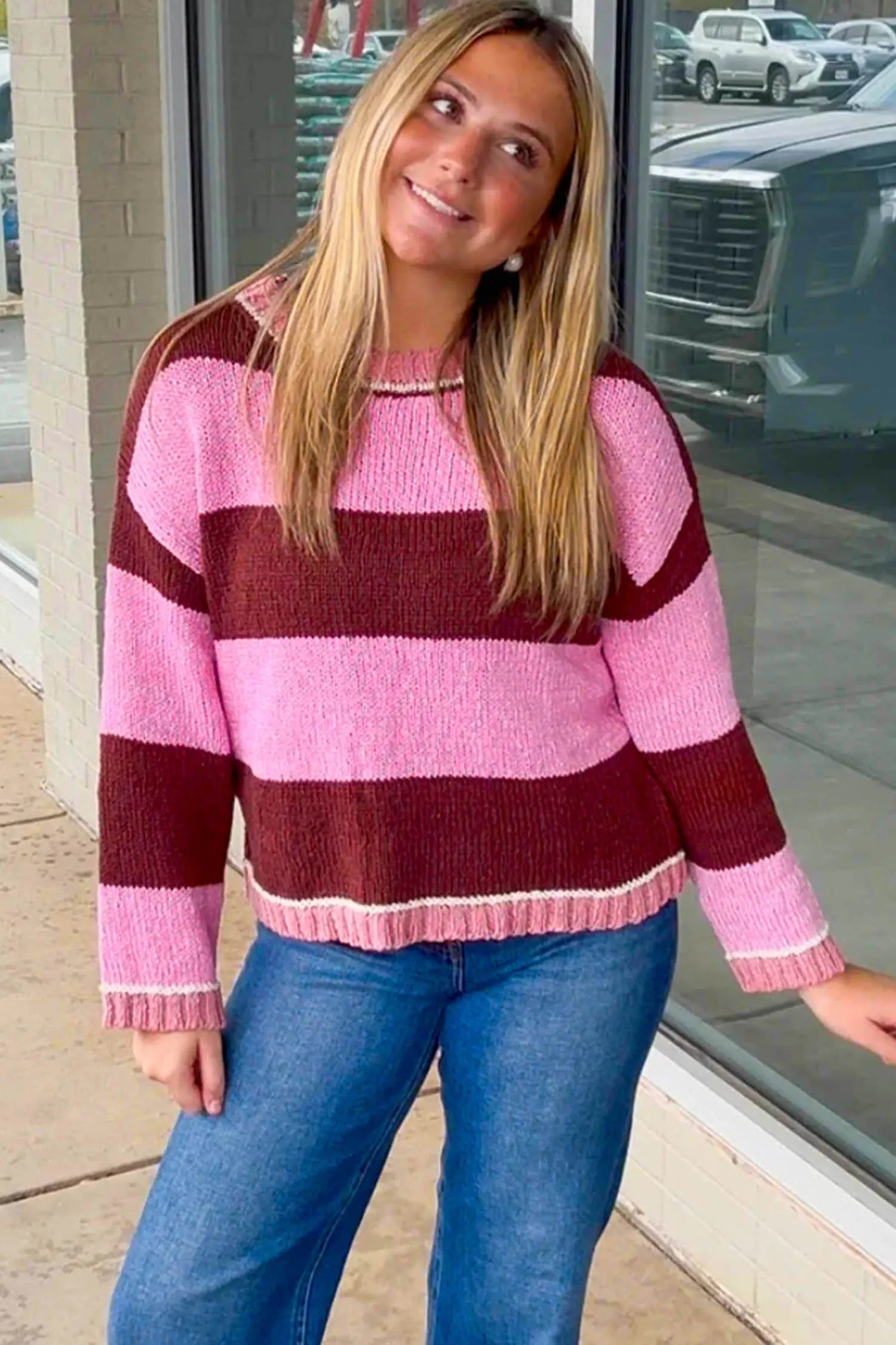 A woman wearing a pink and burgundy striped knit sweater with blue jeans, standing in front of a storefront window.
