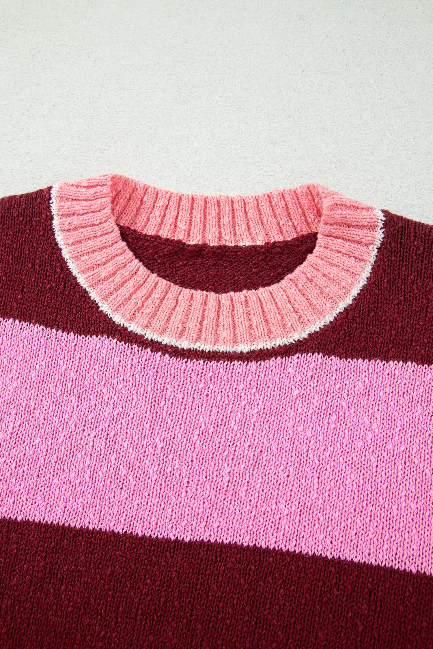 Close-up of a striped knit sweater showing a ribbed crew neck in light pink with a white edge, layered over dark burgundy and a wider medium pink stripe.