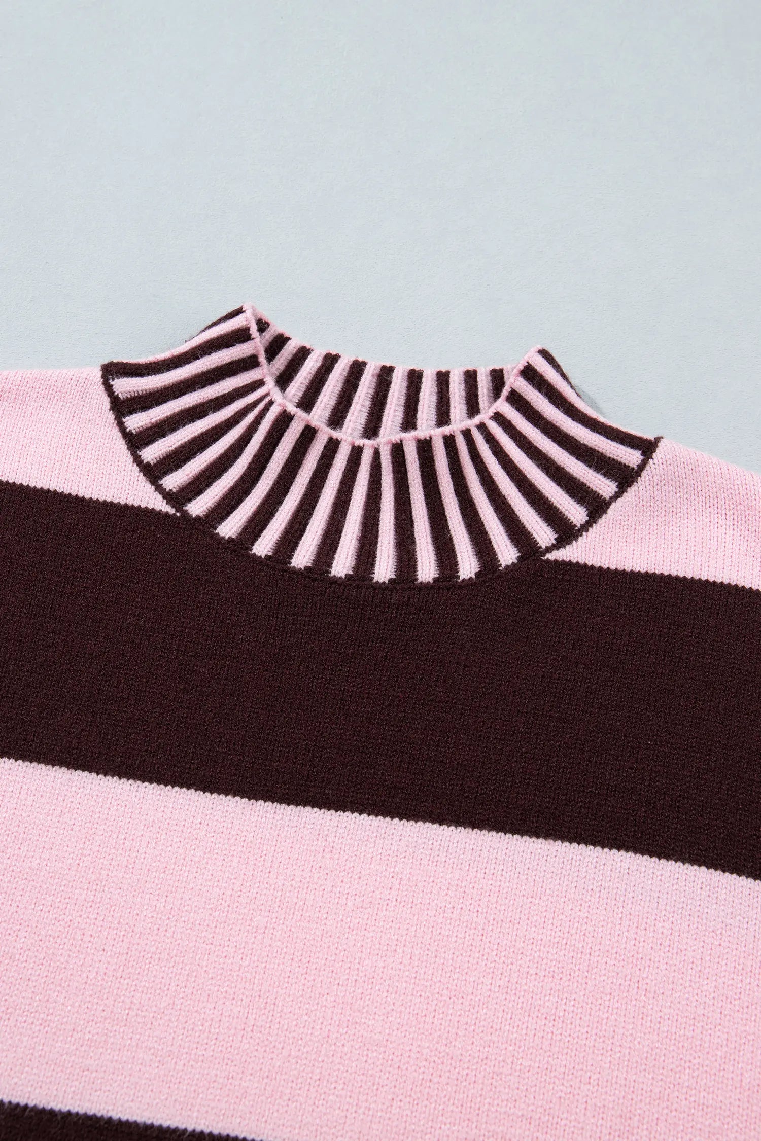 A close-up of a knit sweater showing a striped mock neck collar with alternating dark brown and light pink stripes, a wide dark brown horizontal stripe, and a light pink stripe below it, set against a pale blue-gray background.