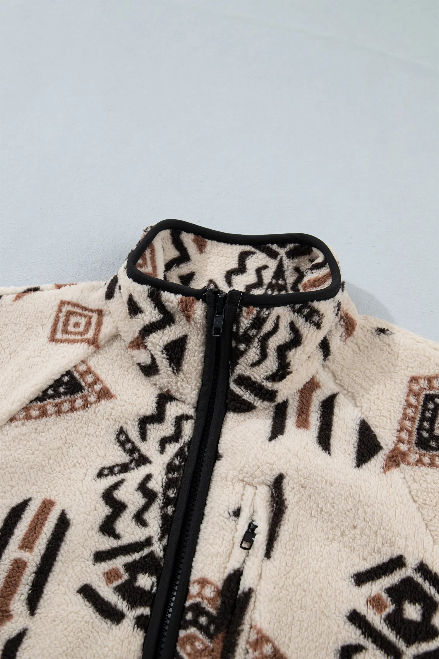 Close-up of a fleece zip-up jacket with a high collar, featuring a cream base with black and brown geometric patterns.