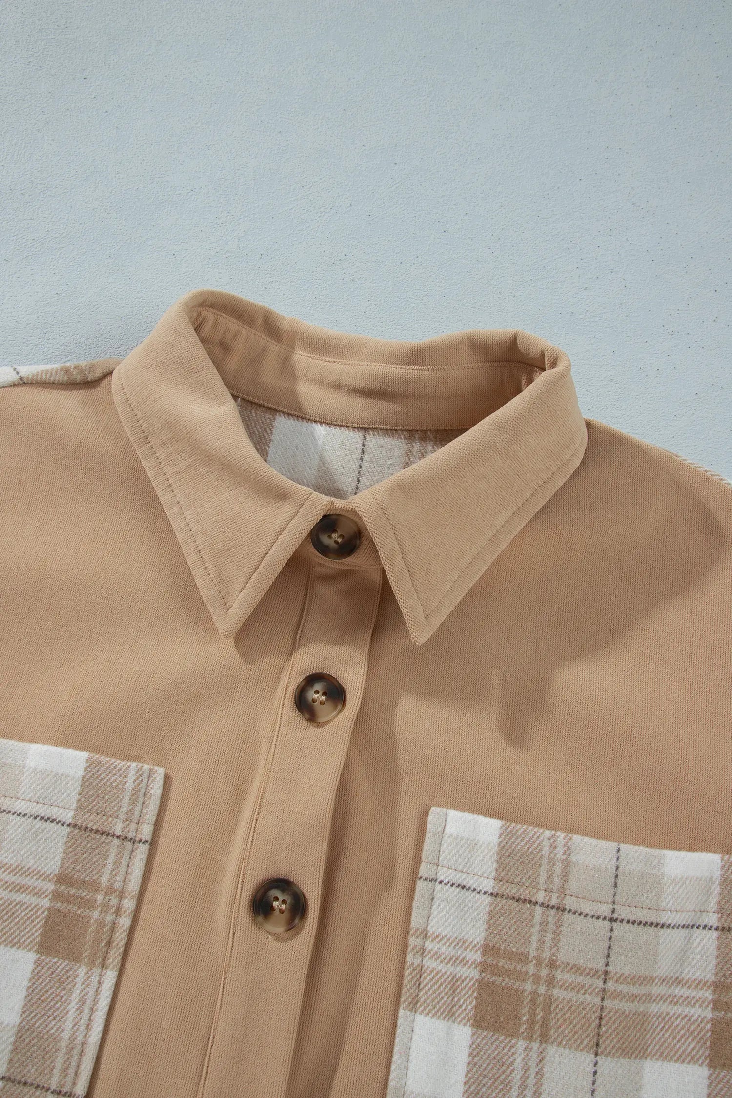 A close-up of a tan button-up shirt with plaid chest pockets and dark brown buttons, laid flat against a light gray background.