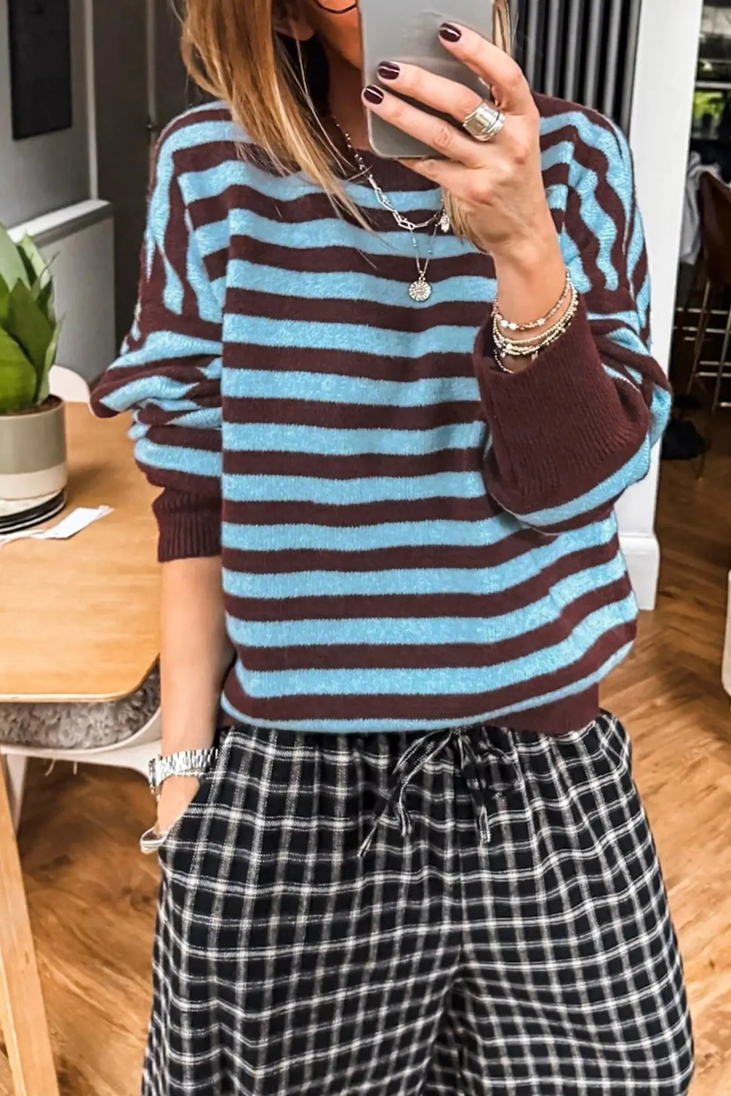 A person wearing a blue and brown horizontally striped knit sweater with dark plaid drawstring pants, taking a mirror selfie in an indoor room.
