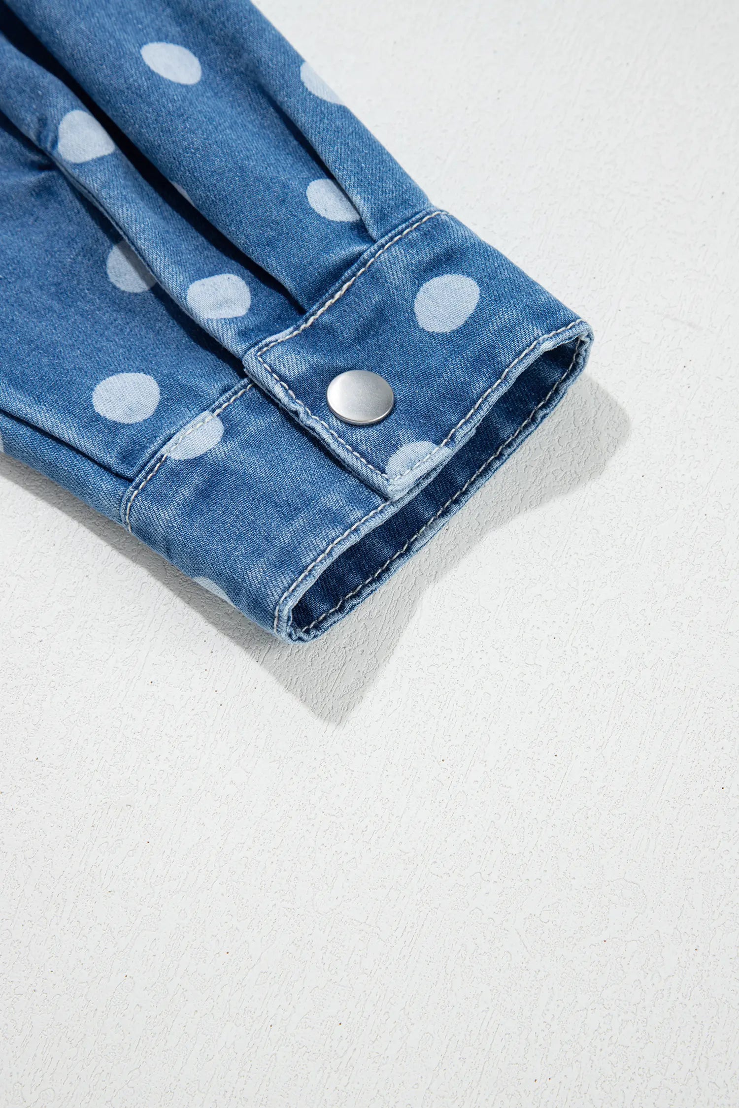 Close-up of blue denim sleeve with large white polka dots and silver snap-button cuff on a light background.