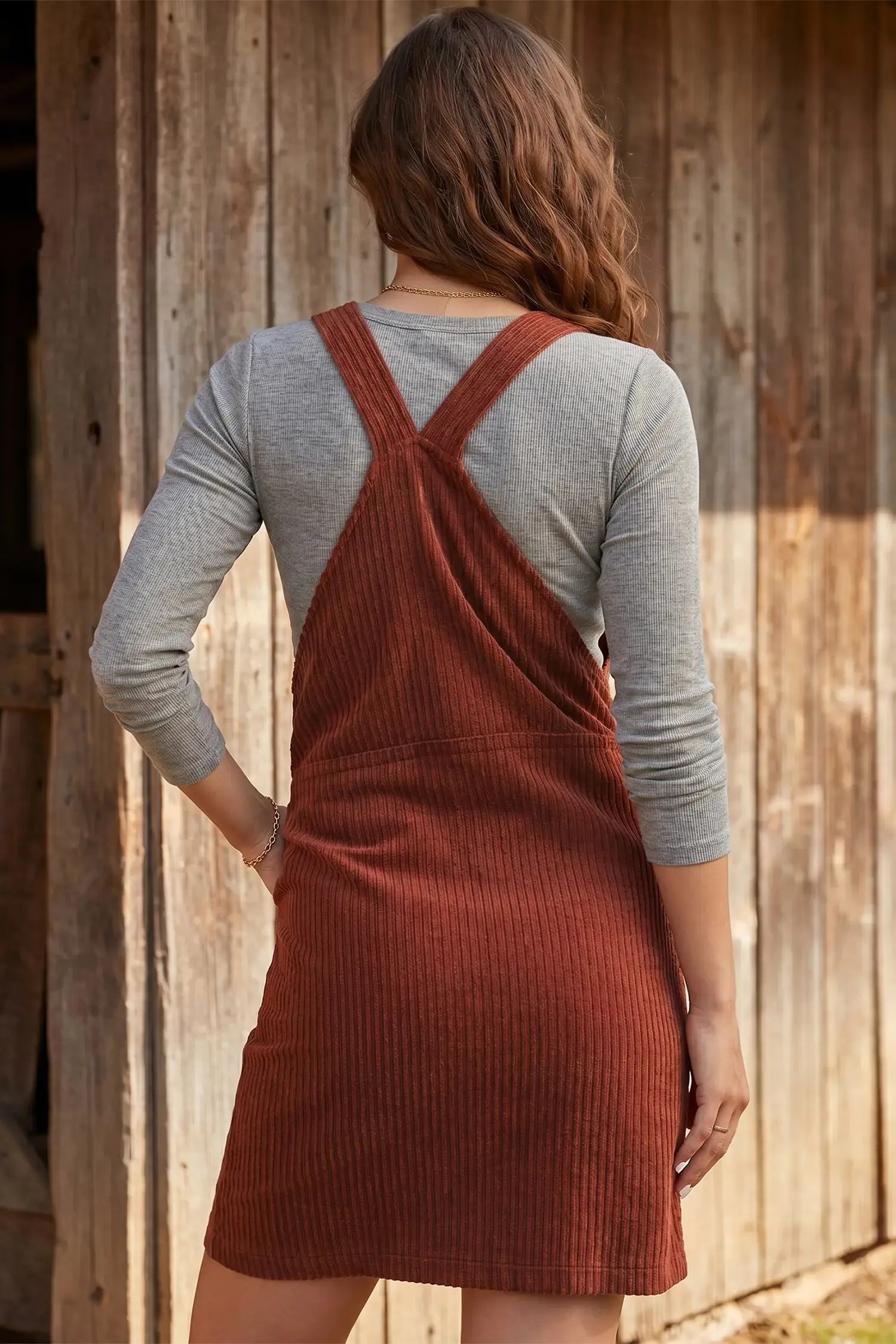A woman viewed from behind wearing a rust-colored ribbed pinafore dress over a heather gray long-sleeve shirt, standing in front of a rustic wooden wall.