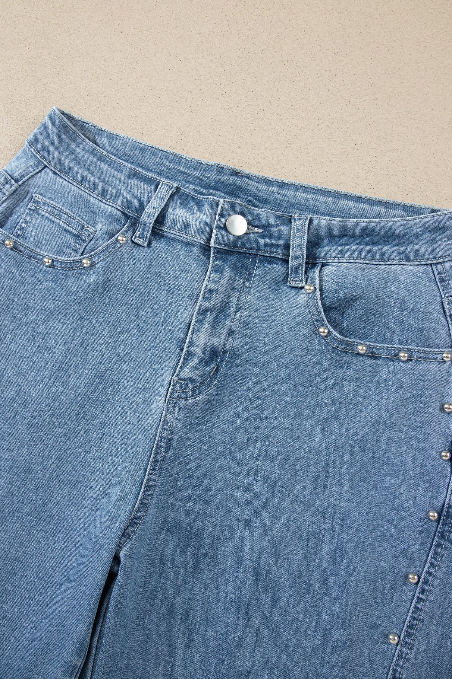 Close-up of a pair of light blue denim jeans with silver metal stud embellishments along the pocket edges, laid flat on a beige background.