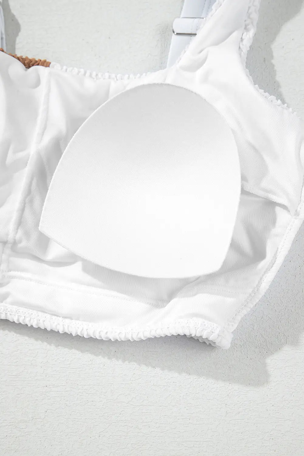 A close-up of a white bikini or bralette top with a smooth, molded white underwire cup and textured ruffled trim along the edge, set against a light gray textured background.