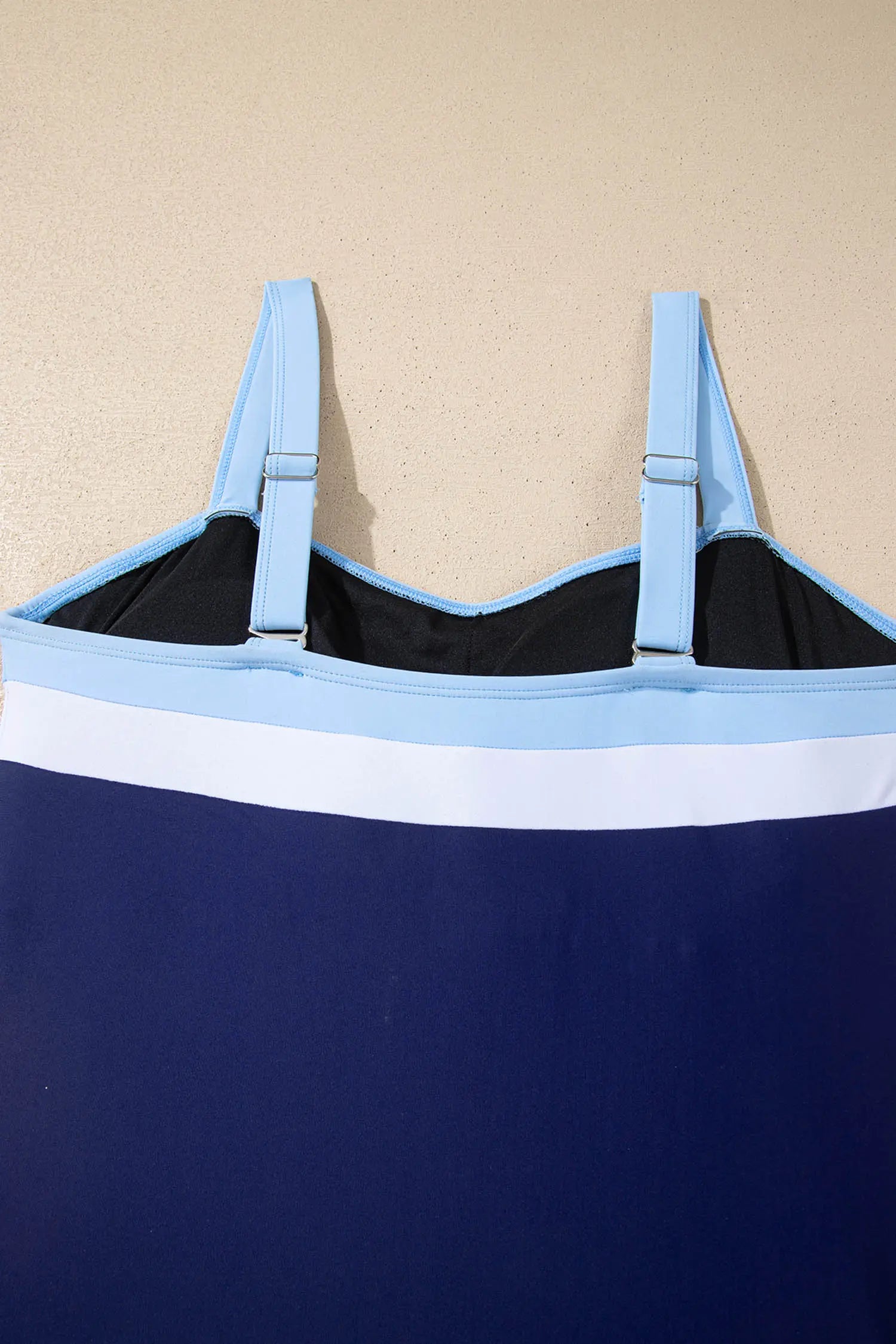 A close-up of a navy blue swimsuit or cover-up with a white horizontal stripe and light blue shoulder straps, shown against a beige textured background.